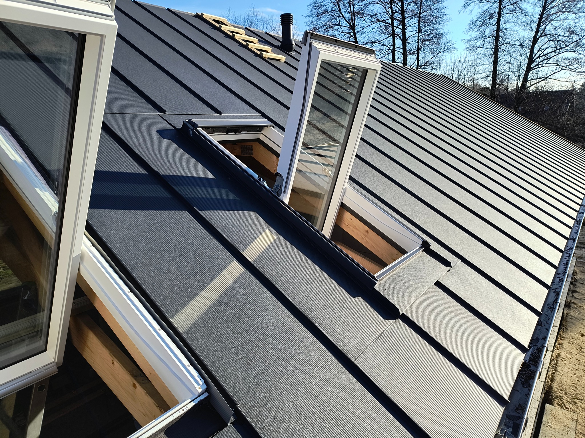 Szary dach z blachy na rąbek stojący z oknami dachowymi, częściowo otwartymi. Widoczne drewniane elementy konstrukcji dachu i fragment nieba.