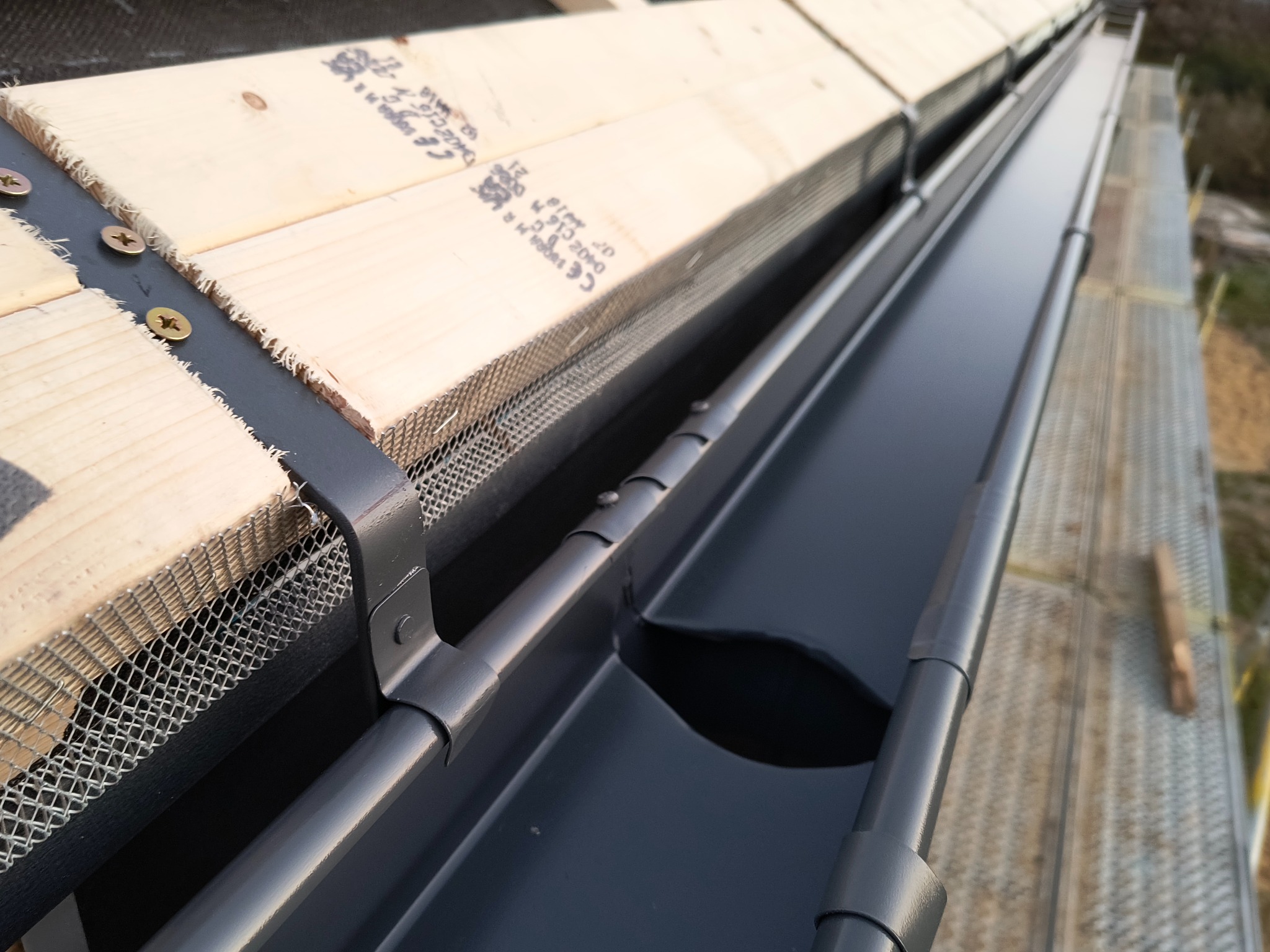 Zamontowana, grafitowa rynna z siatką zabezpieczającą przed liśćmi, przymocowana do drewnianej konstrukcji dachu. Widok z góry, z bliska, z widocznymi detalami mocowania.