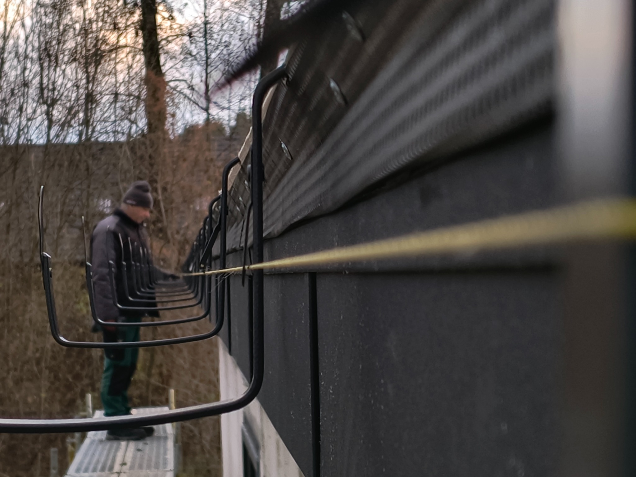 Montaż haków na dachu z blachy trapezowej przez dekarza na rusztowaniu. Widok z dołu, z żółtą linką poziomującą. W tle drzewa bez liści.