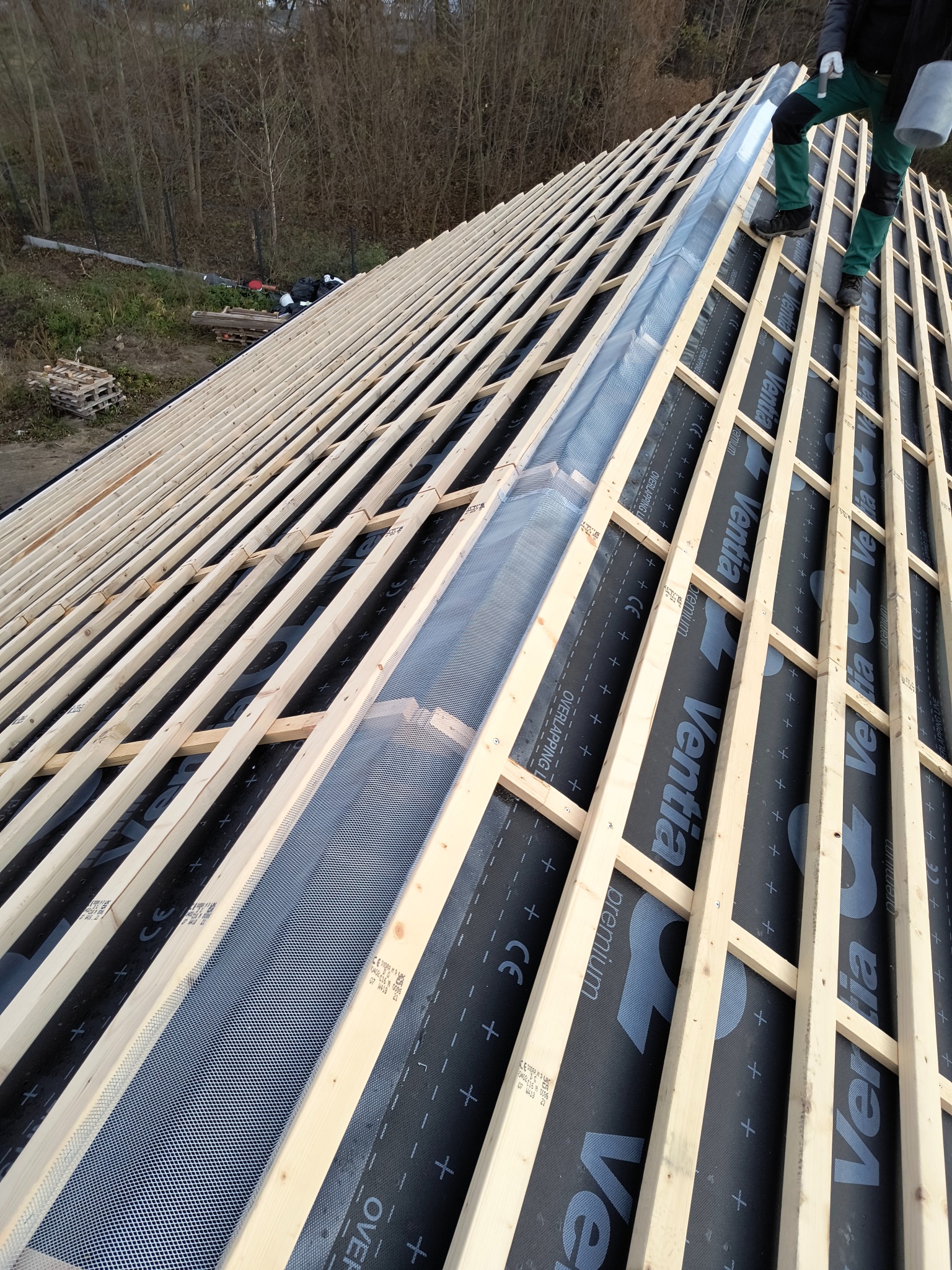 Konstrukcja dachu w trakcie budowy: drewniane łaty, membrana dachowa z nadrukiem 'Ventia Premium', widoczny fragment orynnowania i osoba w zielonych spodniach.