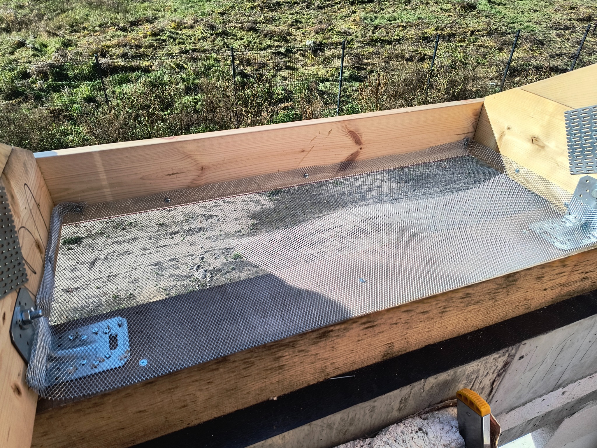 Montaż siatki zabezpieczającej w drewnianej konstrukcji dachu, widoczne metalowe łączniki i narzędzie. Tło stanowi trawa i ogrodzenie.