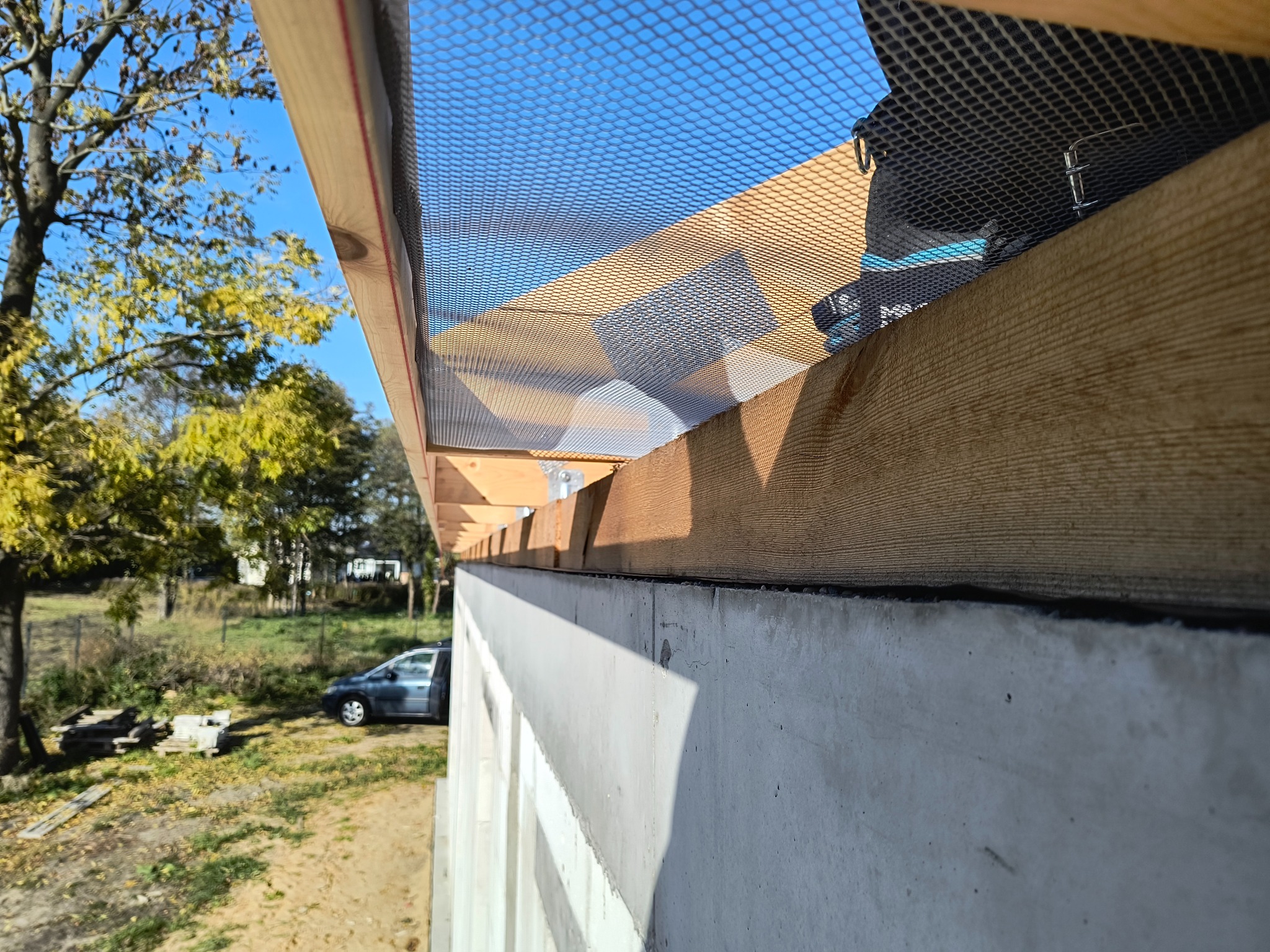 Montaż siatki okapowej na drewnianej konstrukcji dachu, widok z dołu na tle błękitnego nieba i zieleni drzew. Detal zabezpieczenia przed ptakami i gryzoniami.