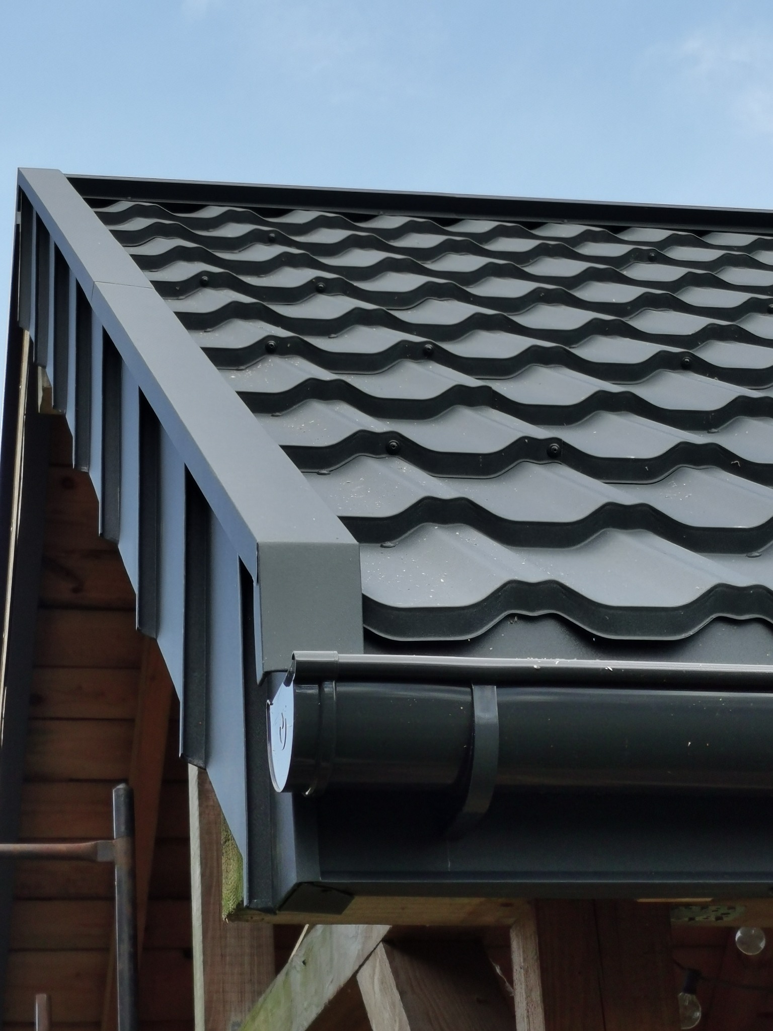Szczegółowe ujęcie fragmentu dachu z szarej blachy z widocznymi obróbkami blacharskimi i systemem rynnowym, na tle błękitnego nieba i drewnianej konstrukcji.