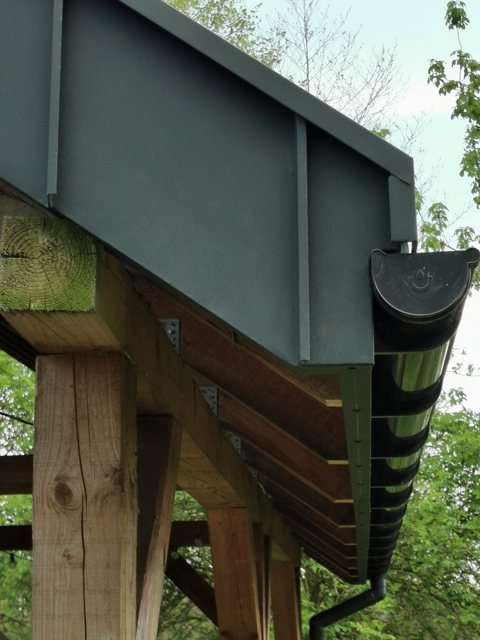 Szczegółowe ujęcie okapu dachu z ciemną blachą, drewnianą konstrukcją wsporczą i systemem rynnowym, na tle zieleni drzew.
