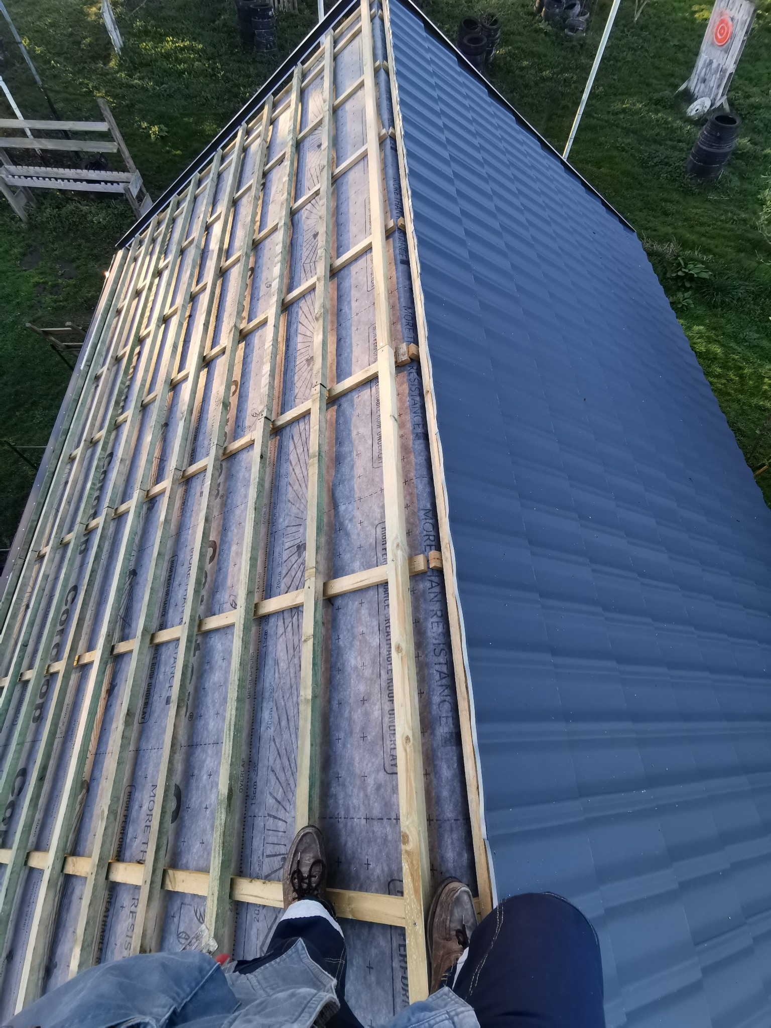 Częściowo pokryty dach z widoczną konstrukcją drewnianą i membraną. Fragment dachu pokryty ciemną blachą. Widok z góry na osobę stojącą na dachu w trakcie prac.