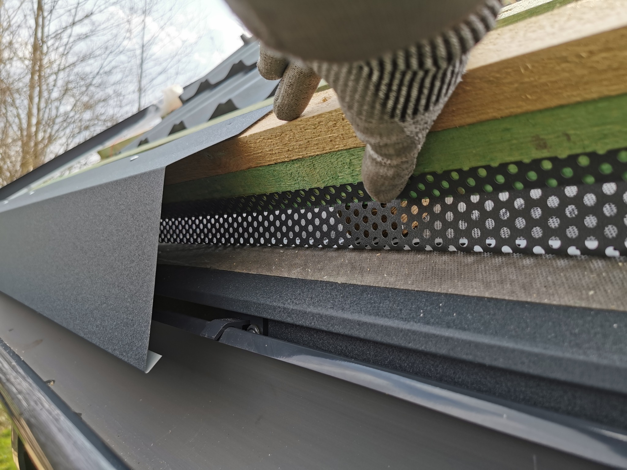 Montaż okapu dachowego z perforowaną listwą wentylacyjną i rynną w kolorze grafitowym. Widoczne warstwy konstrukcji dachu i dłoń w rękawicy roboczej.