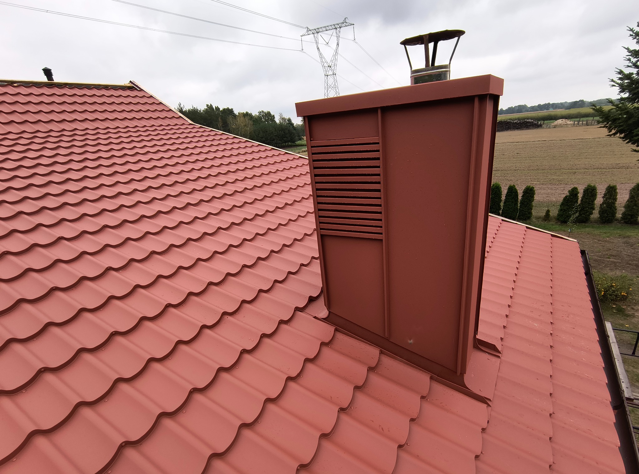 Czerwona dachówka ceramiczna na skośnym dachu domu jednorodzinnego, z kominem wentylacyjnym w kolorze dachówki. W tle słup wysokiego napięcia i zielone pola.