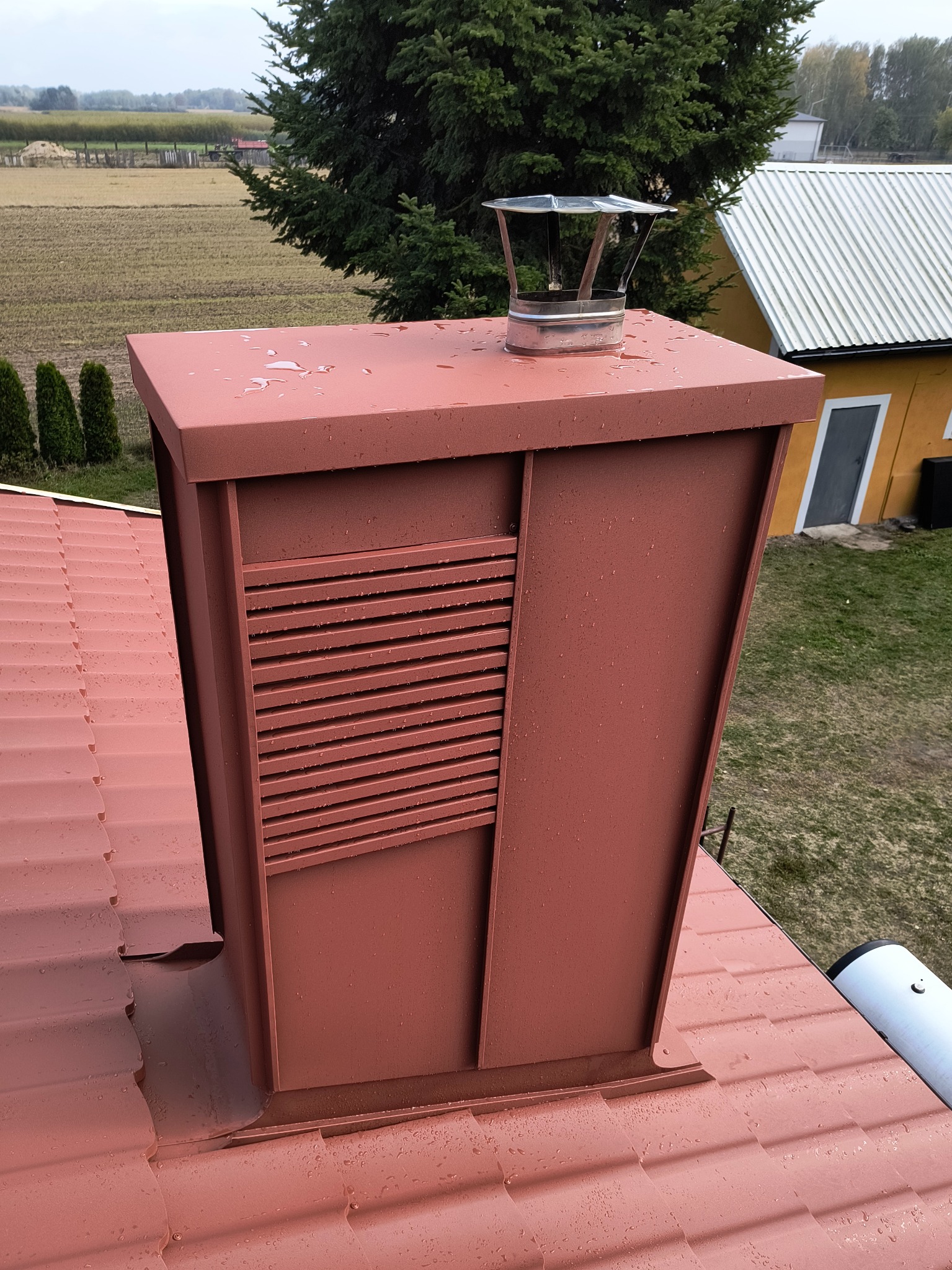 Czerwony komin wentylacyjny z metalowym daszkiem na dachu z dachówki ceramicznej, widok z góry, krople deszczu na powierzchni, w tle budynek i drzewa.