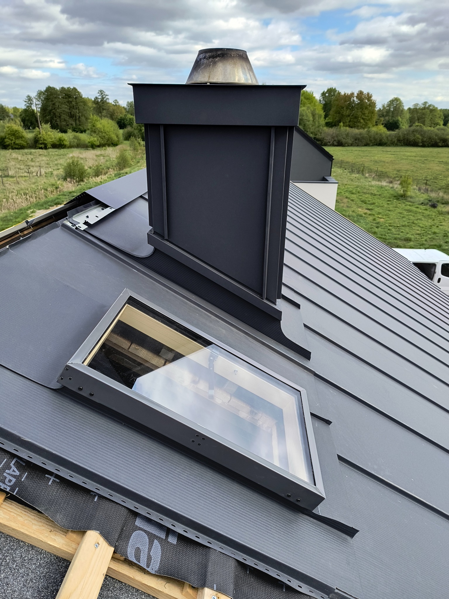 Szary dach z blachy na rąbek stojący, z oknem dachowym i kominem. Widoczne detale obróbek blacharskich i elementy montażowe. W tle zieleń drzew i łąk.