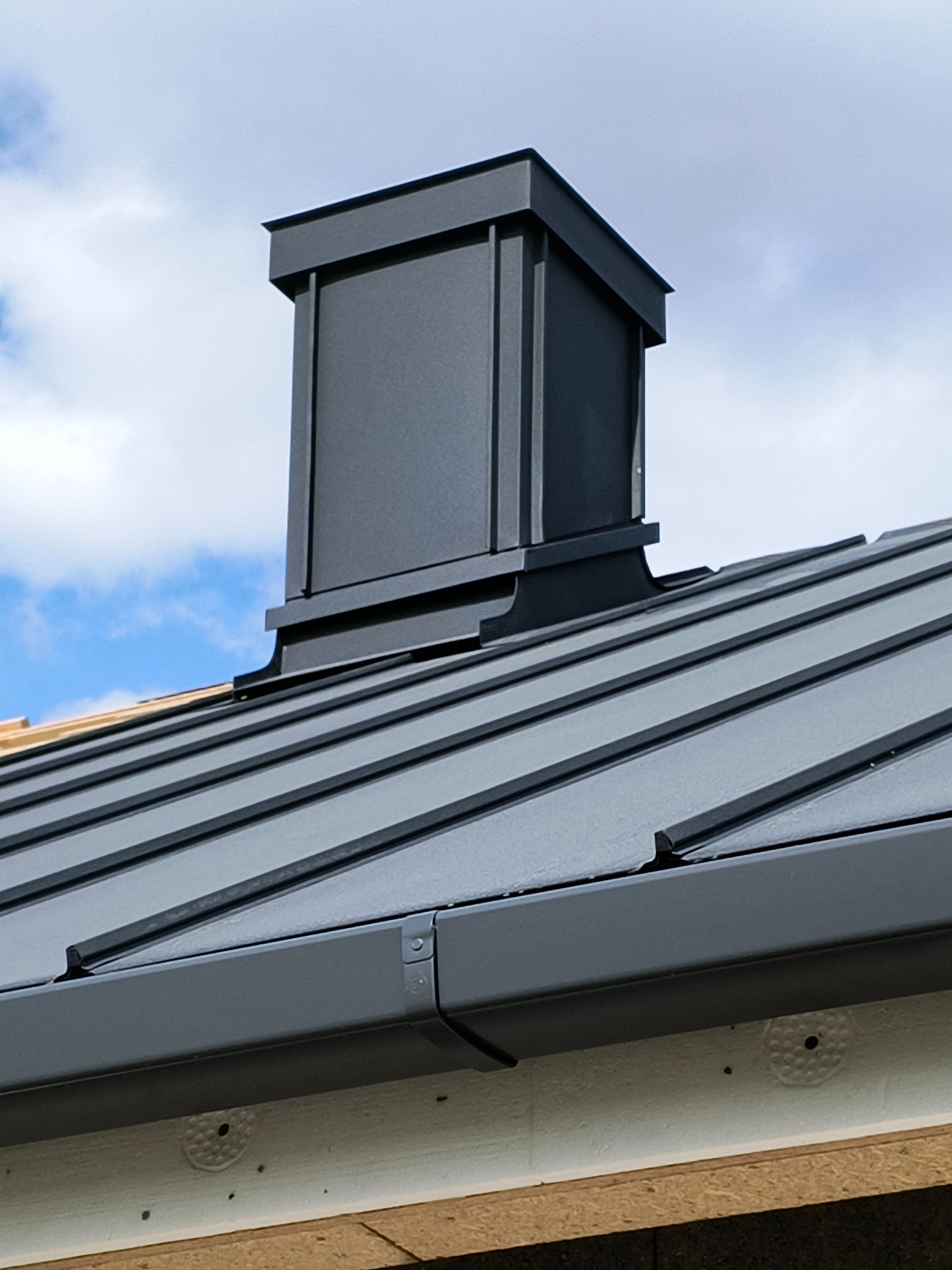 Szary dach z blachy trapezowej i komin z blachy na tle błękitnego nieba z chmurami. Widoczne detale obróbki blacharskiej i mocowania.