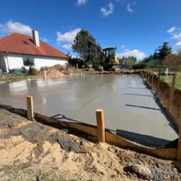 Wylany fundament pod budynek jednorodzinny, szalunek z desek, widoczny mały żółty koparka na tle błękitnego nieba i domu z czerwoną dachówką.