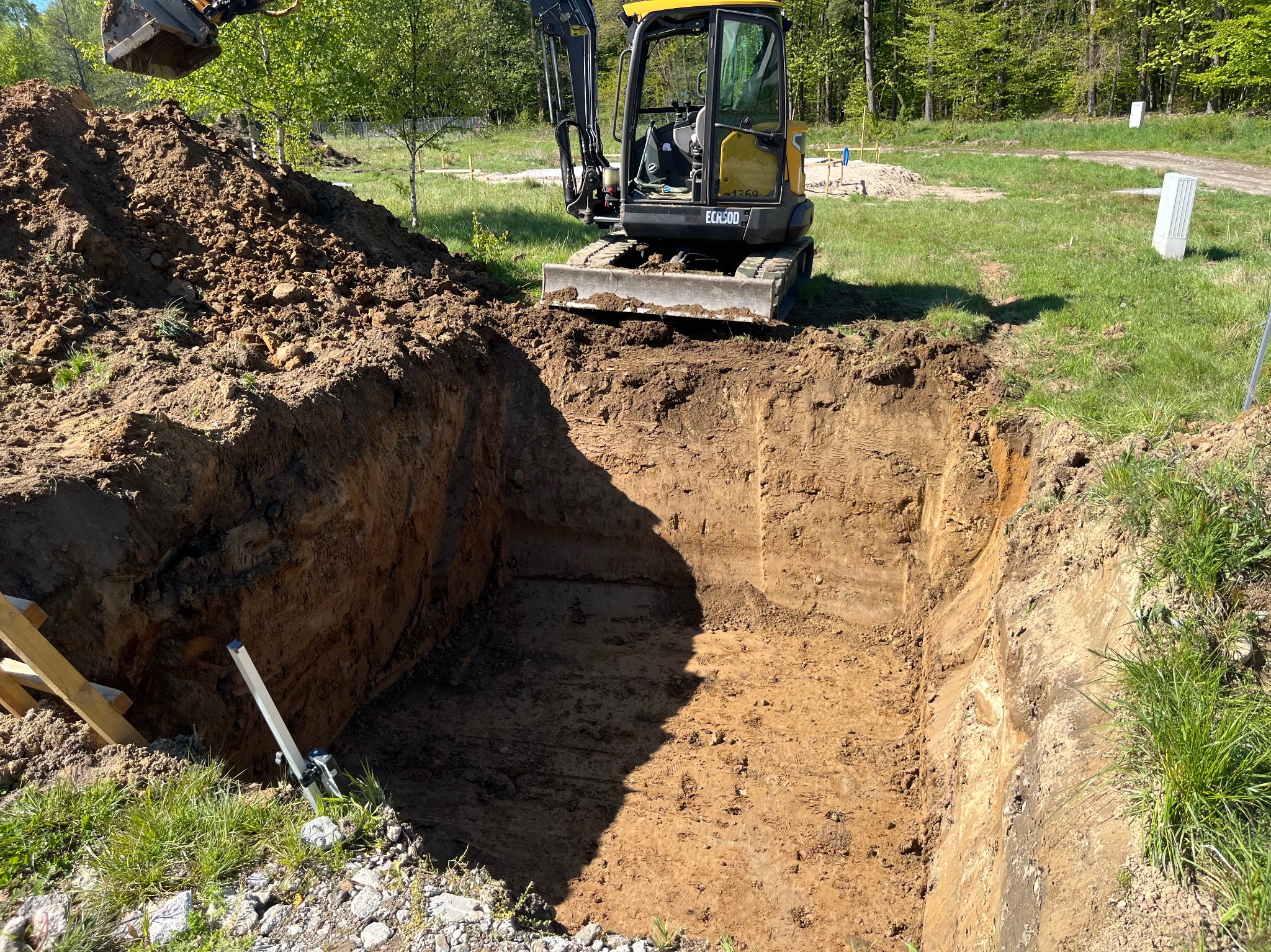 Żółta koparka przy wykopie w ziemi, obok sterta ziemi i poziomica. Widoczne drzewa w tle, słoneczny dzień. Prace ziemne w toku.