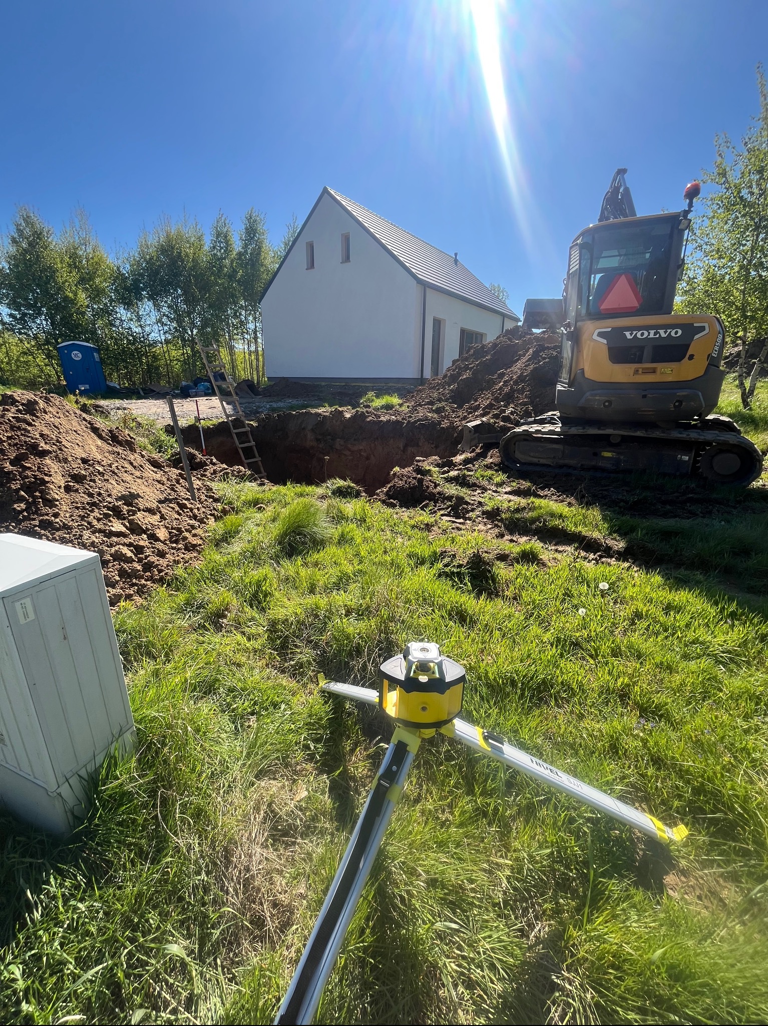 Żółta koparka Volvo przy wykopie obok domu, z niwelatorem na pierwszym planie. Wykop zabezpieczony drabiną, widoczna przenośna toaleta.