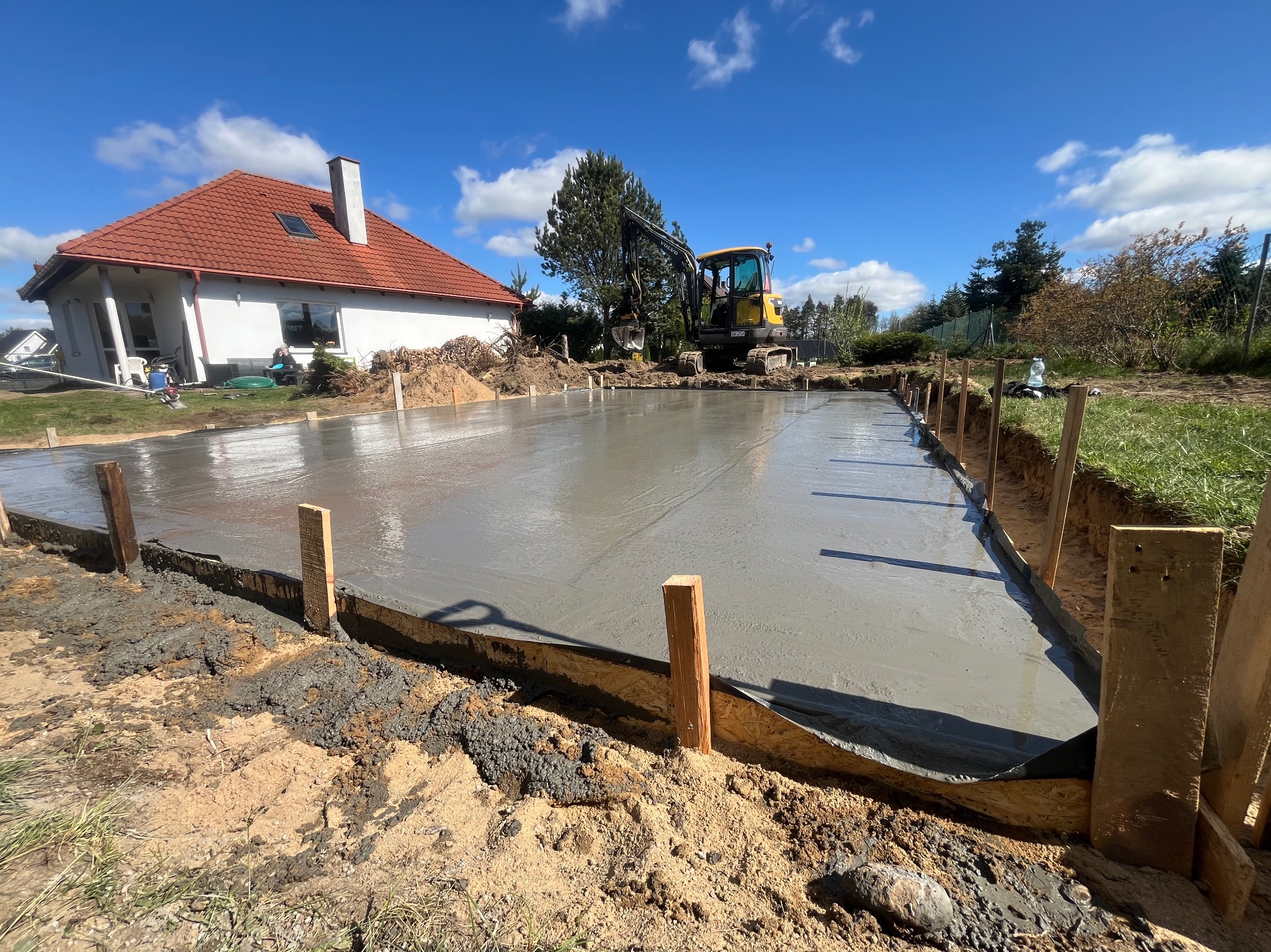 Wylany fundament pod budynek jednorodzinny, szalunek z desek, widoczny mały żółty koparka na tle błękitnego nieba i domu z czerwoną dachówką.