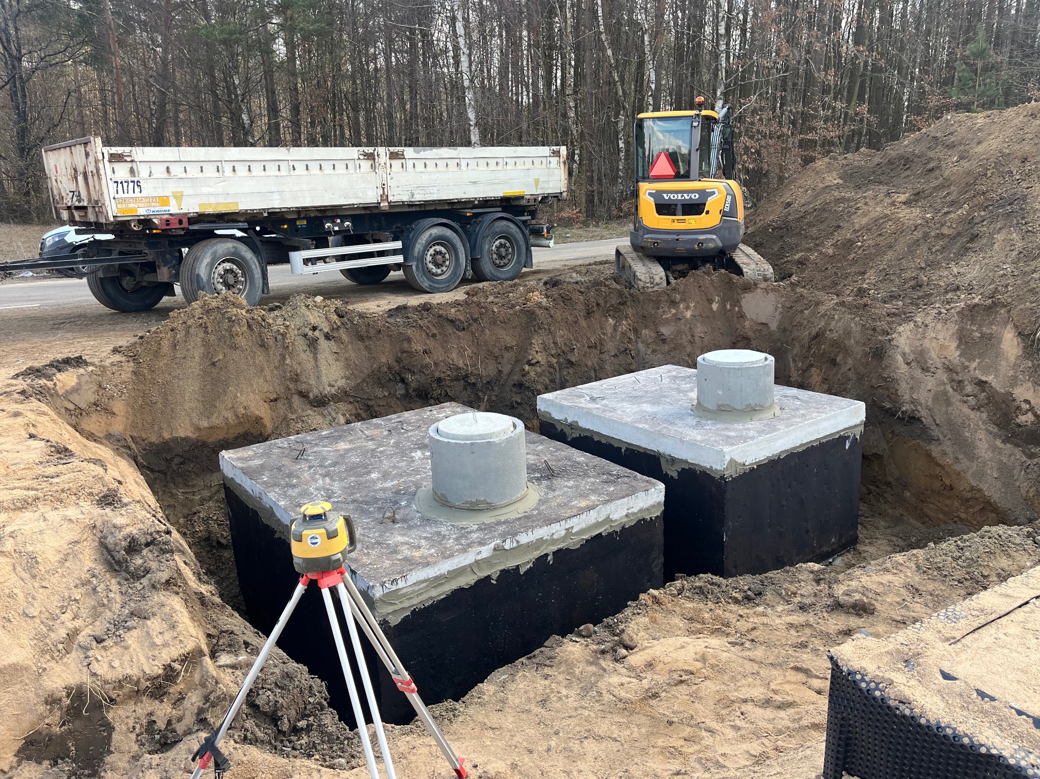 Dwa betonowe szamba w wykopie, żółta koparka Volvo i biała ciężarówka w tle. Na pierwszym planie niwelator laserowy na statywie. Prace ziemne w toku.