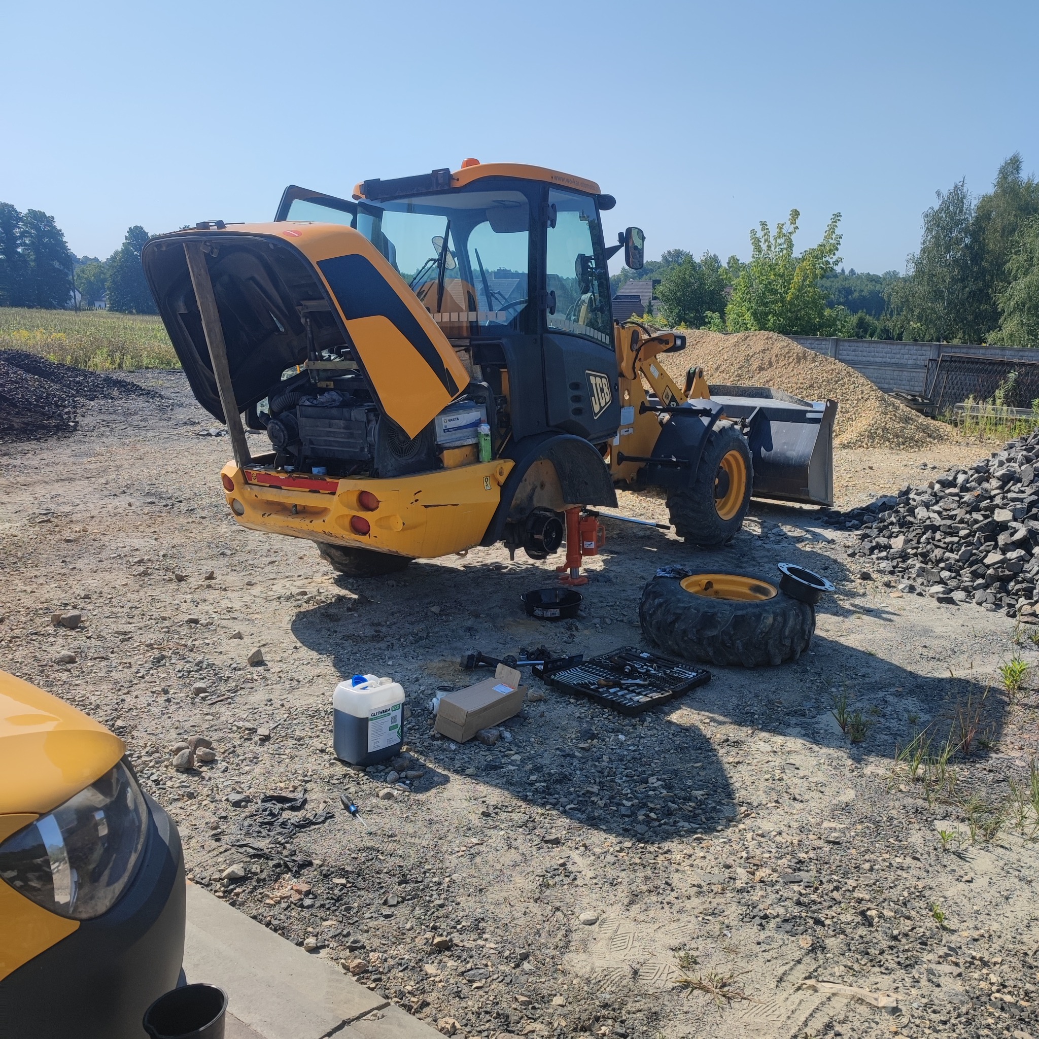 Żółta koparko-ładowarka JCB z otwartą maską silnika i zdjętym kołem, podnośnik, narzędzia i płyny eksploatacyjne obok, teren budowy w tle.