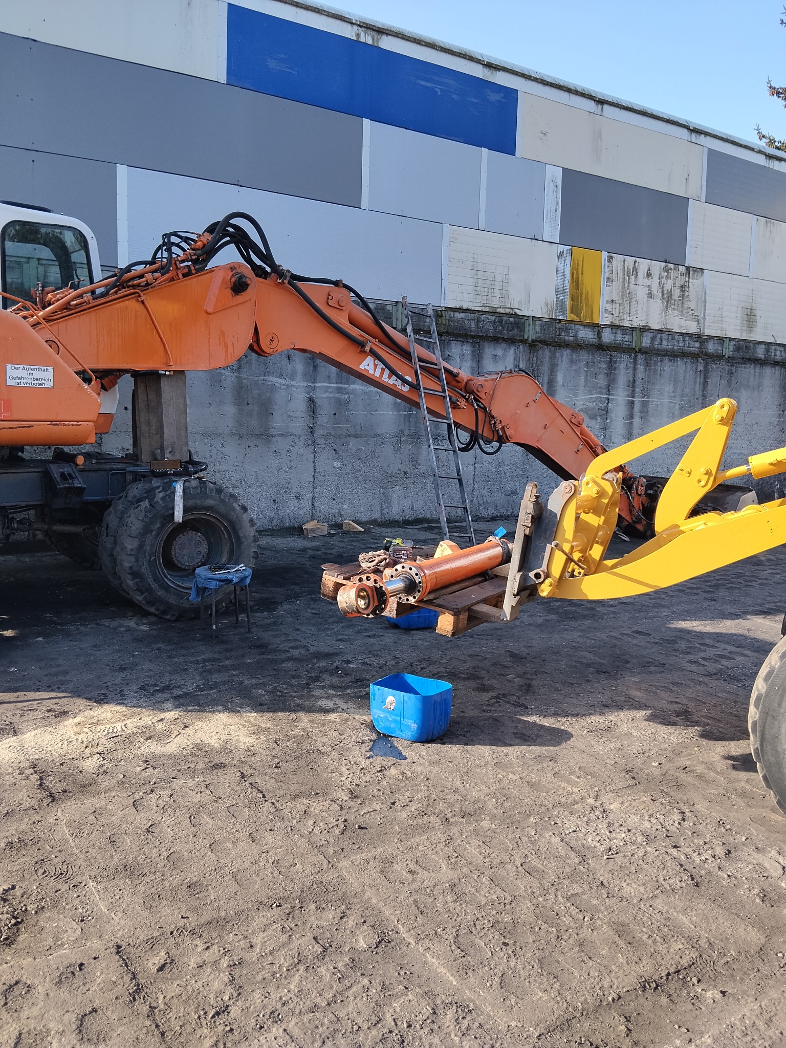 Pomarańczowa koparka Atlas z odłączonym elementem roboczym, podpartym żółtym ramieniem. Obok narzędzia i niebieski pojemnik na ziemistym podłożu, drabina oparta o ramię koparki.