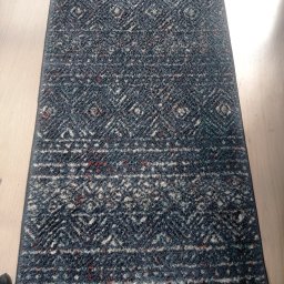 Czyste Meble Alex - Wzorzysty, prostokątny dywan w odcieniach szarości i bieli z akcentami czerwieni, leżący na jasnej drewnianej podłodze. Widoczne delikatne cienie i struktura materiału.