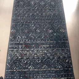 Wzorzysty, prostokątny dywan w odcieniach szarości i bieli z akcentami czerwieni, leżący na jasnej drewnianej podłodze. Widoczne delikatne cienie i struktura materiału.