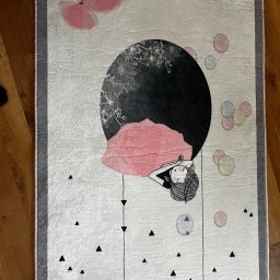 Czyste Meble Alex - Wzorzysty dywan z motywem leżącej postaci i abstrakcyjnymi elementami na drewnianej podłodze, widziany z góry. Szaro-biała kolorystyka z akcentami różu i czerni.