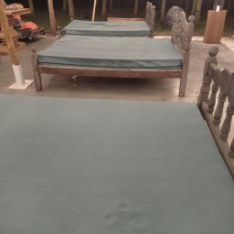 Czyste Meble Alex - Ustawione na zewnątrz łóżka z szarymi materacami, oświetlone girlandami, w tle drzewa i trawa. Materace wyglądają jakby wymagały odświeżenia.