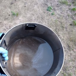 Czyste Meble Alex - Brudna woda po czyszczeniu w zbiorniku odkurzacza. Widoczna ręka w niebieskiej rękawicy. Tło to ziemia z fragmentami trawy. Ujęcie z góry.