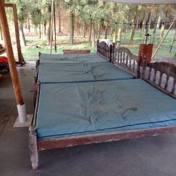 Czyste Meble Alex - Długi, drewniany tapczan z niebieskim materacem pod namiotem, na tle drzew. Widoczne ślady użytkowania na materacu. Dekoracyjne oświetlenie na górze.