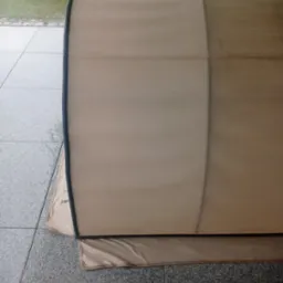 Fragment beżowej, zwiniętej markizy na szarym, kamiennym podłożu. Widoczne ślady czyszczenia, różnica w odcieniu. Trawa i drzewo w tle.