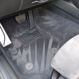 Czyste Meble Alex - Wnętrze auta po czyszczeniu: świeże ślady po praniu na dywaniku pod pedałami i w okolicy fotela. Ciemna tapicerka, widoczne mocowania dywanika.