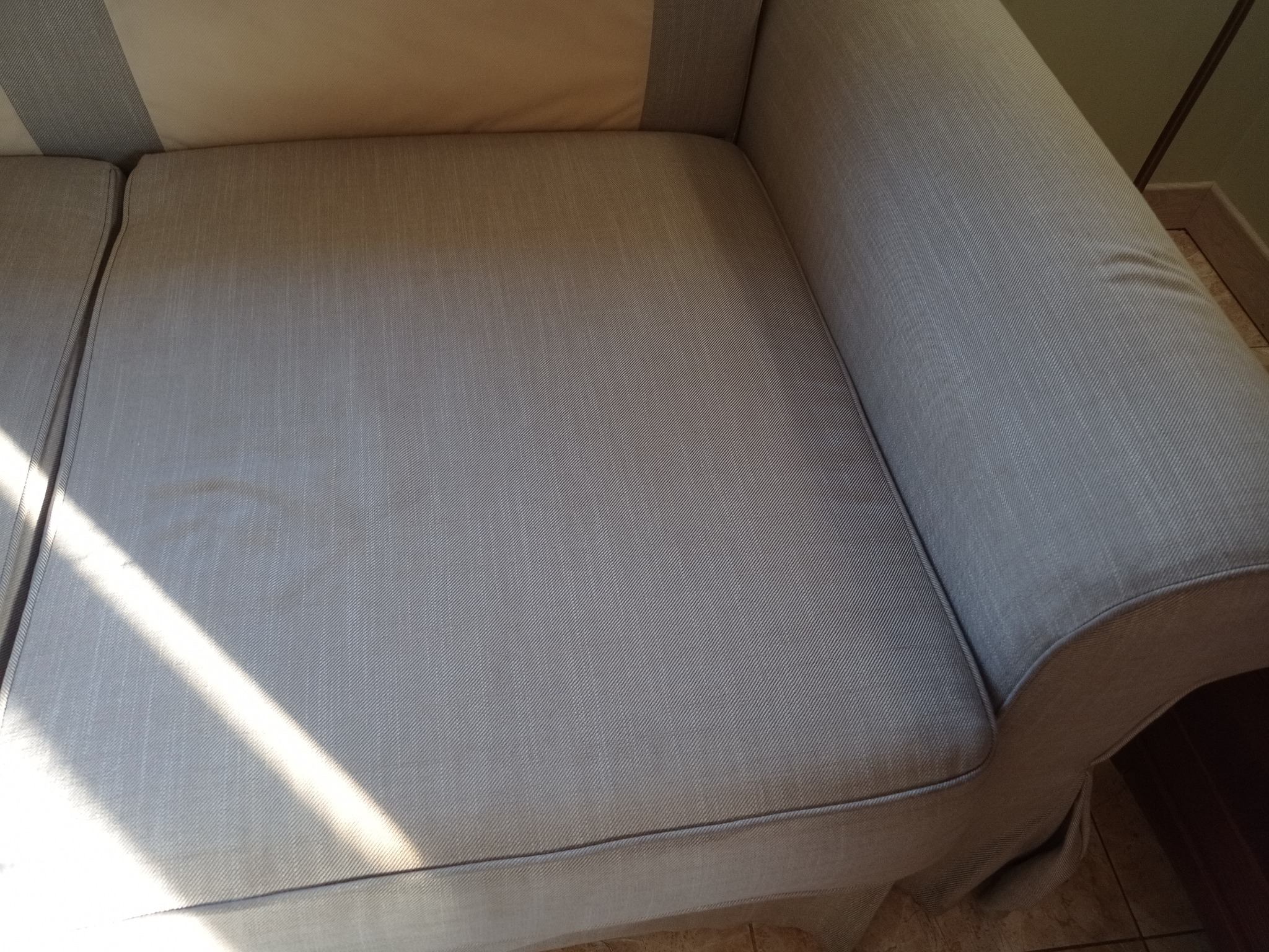 Jasnoszara kanapa z widocznym siedziskiem i podłokietnikiem, oświetlona promieniami słonecznymi. Widoczna faktura materiału i fragment podłogi.