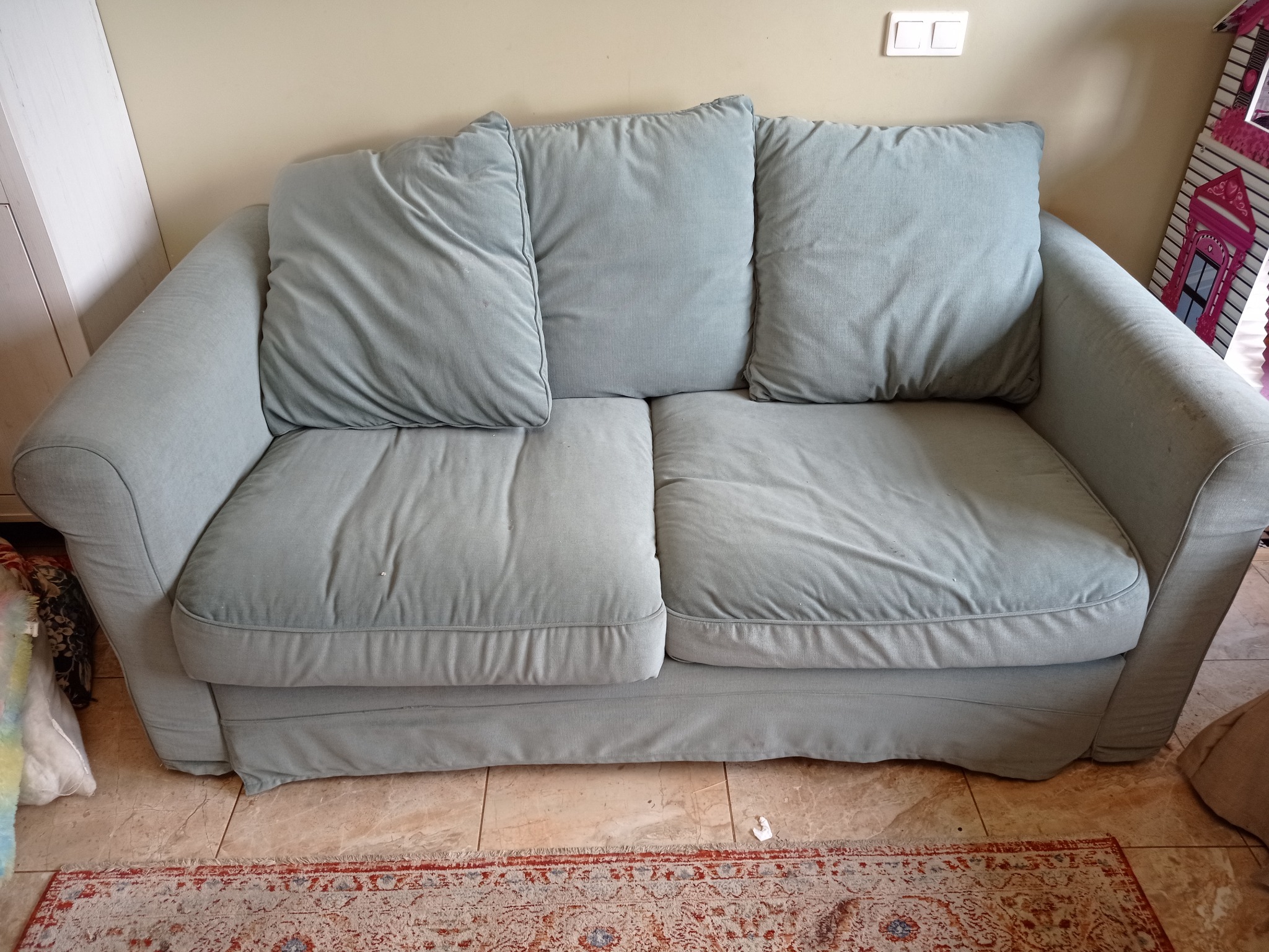 Jasnoszara sofa z trzema poduszkami na tle beżowej ściany i wzorzystego dywanu. Widoczne gniazdko elektryczne i fragment dziecięcej zabawki.