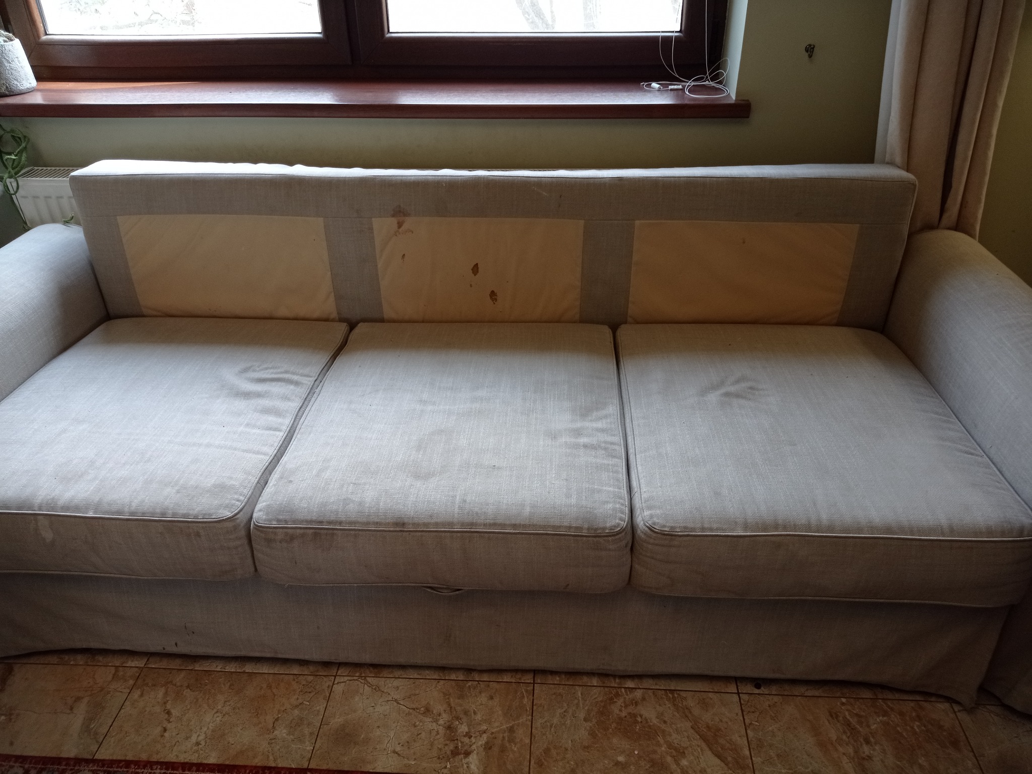 Szara, trzysiedziskowa kanapa z plamami na oparciu, stojąca na brązowej podłodze, z oknem w tle. Widoczne ślady użytkowania.