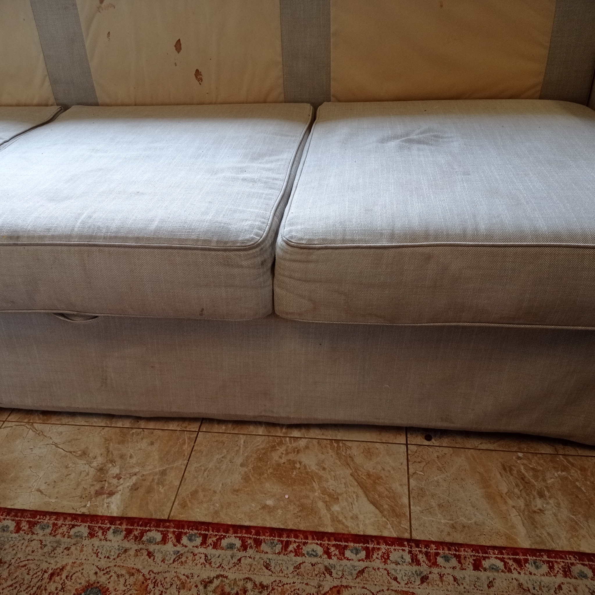 Fragment sofy z jasną tapicerką, widoczne plamy i zabrudzenia na siedzisku. W tle wzorzysta tkanina, na dole brązowe płytki podłogowe i fragment dywanu.