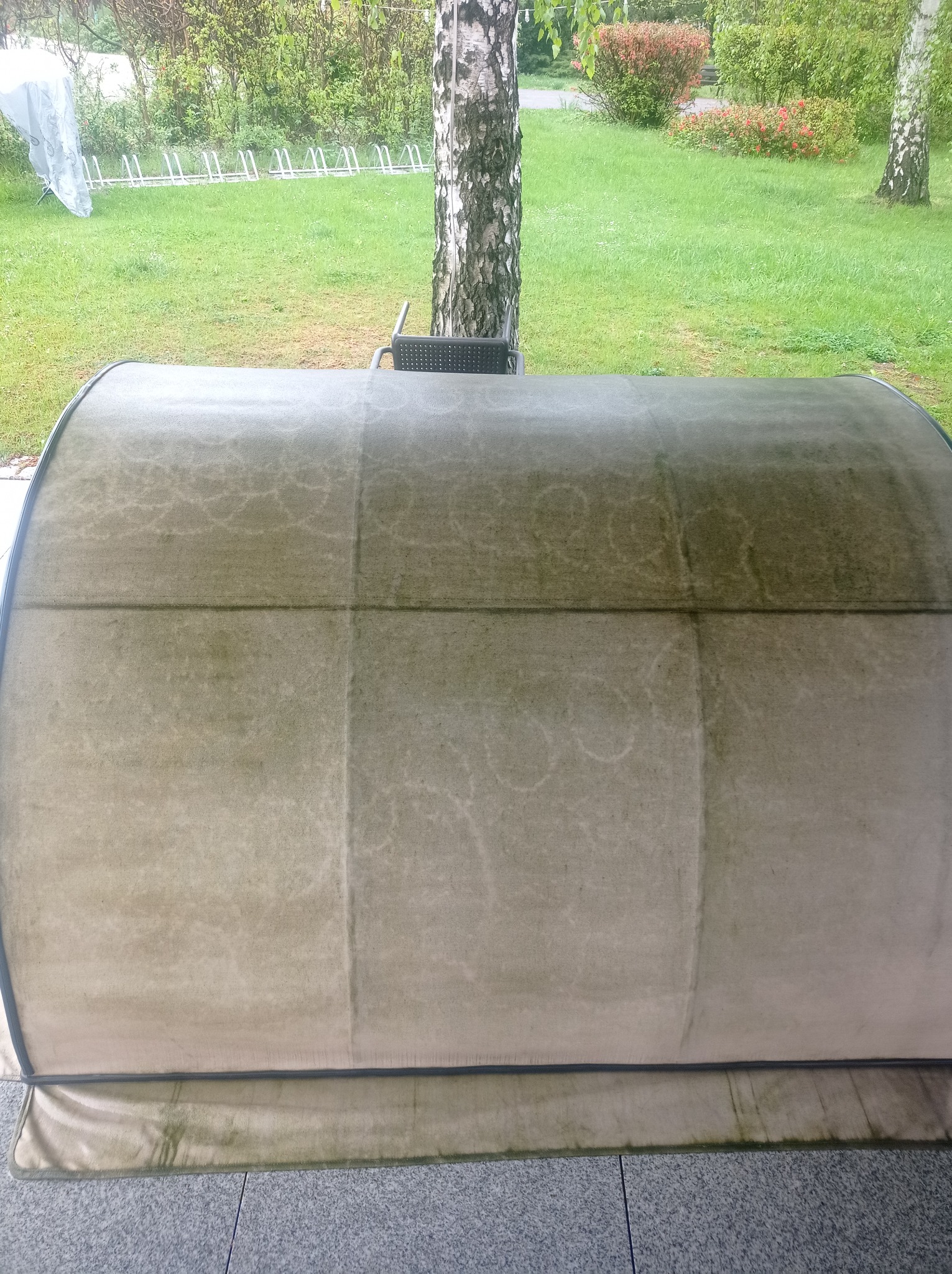 Widok z bliska na czyszczoną, beżową kanapę ogrodową z widoczną różnicą w czystości po lewej stronie. Tło stanowi trawnik i drzewa.