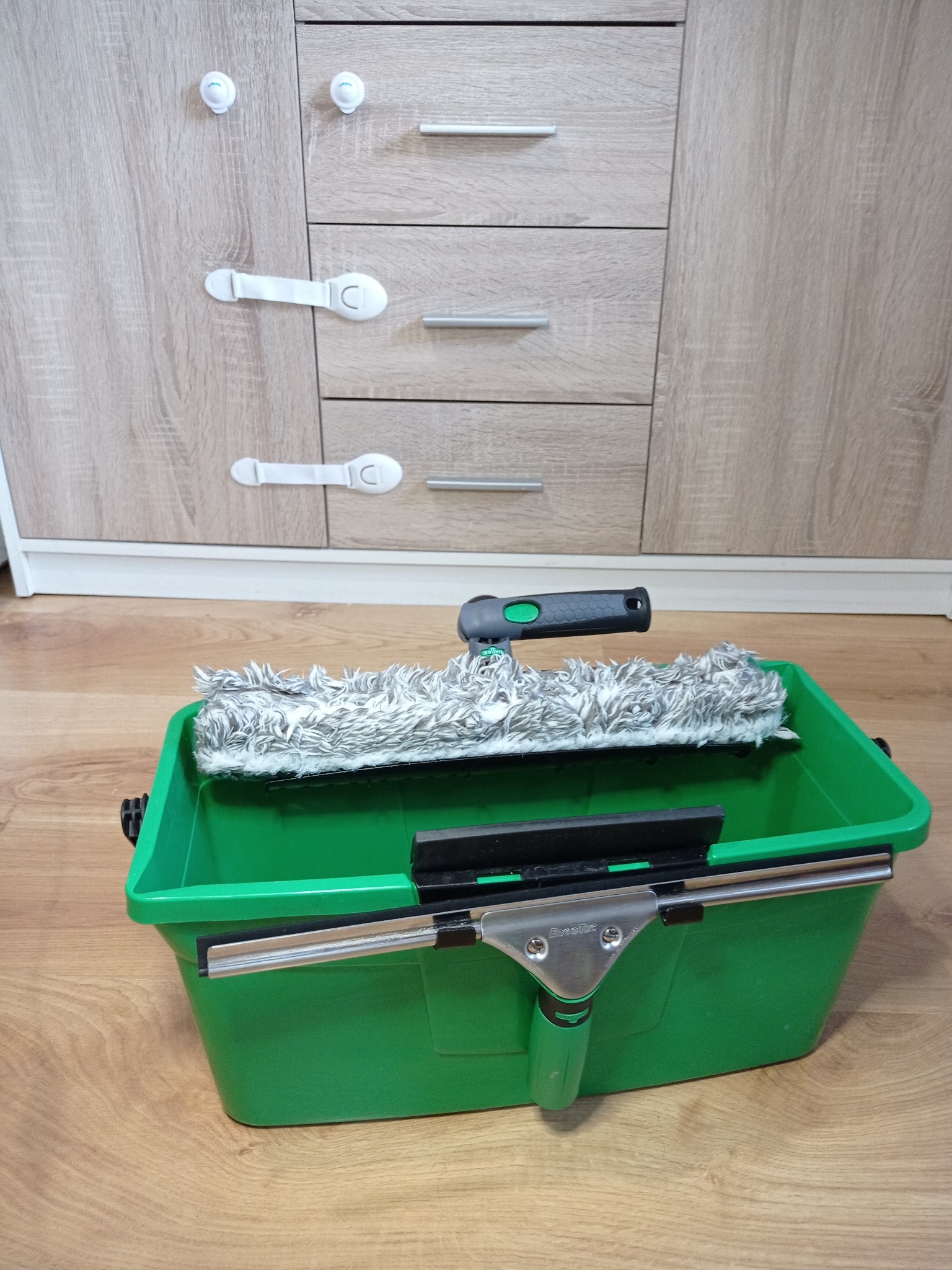 Zielony pojemnik z mopem do mycia okien i ściągaczką na drewnianej podłodze, na tle szafki z zabezpieczeniami na szufladach.