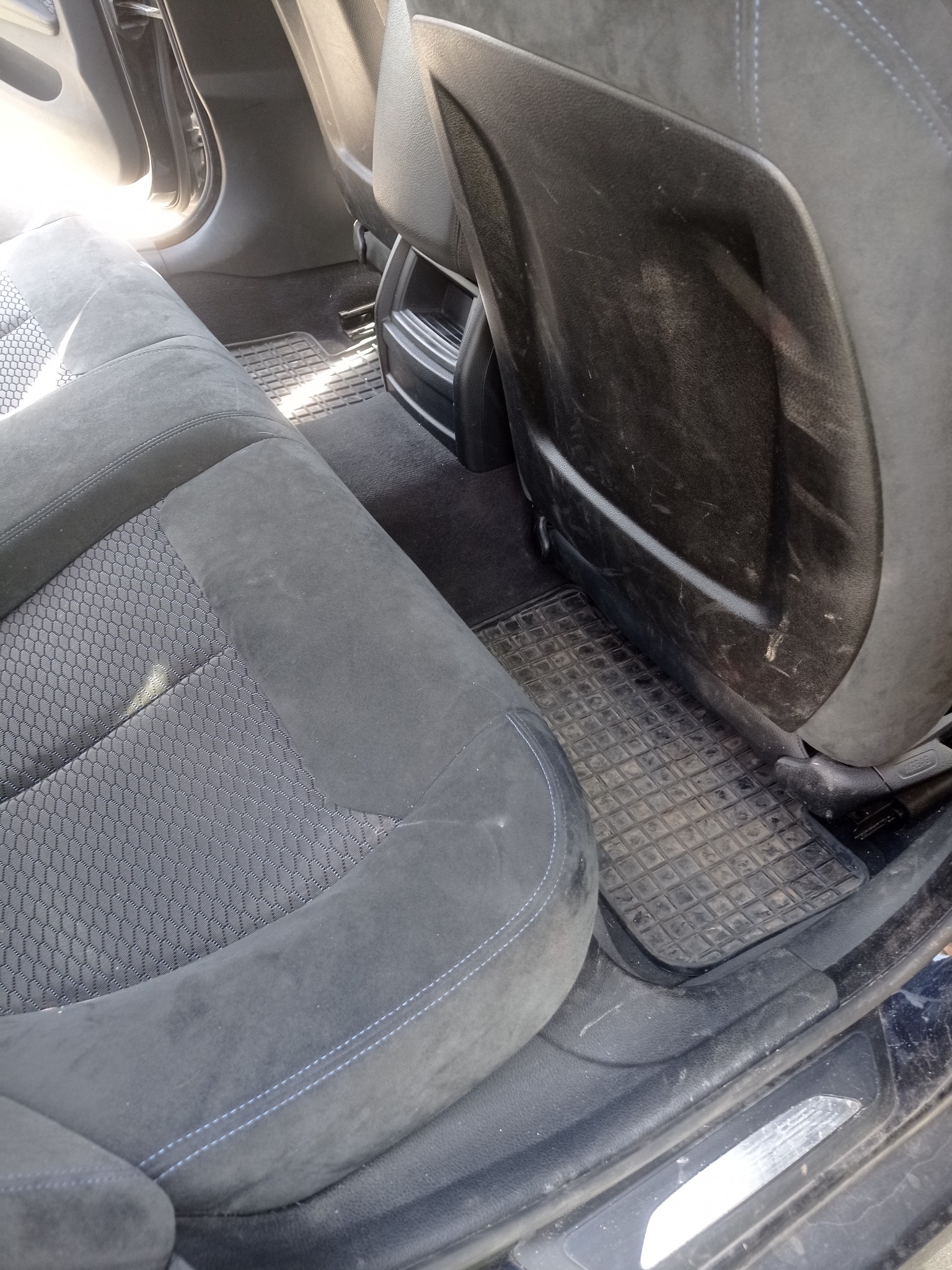 Zabrudzone tylne siedzenie samochodu z czarną tapicerką, widoczne plamy na oparciu fotela i gumowe dywaniki. Wnętrze auta wymaga czyszczenia.