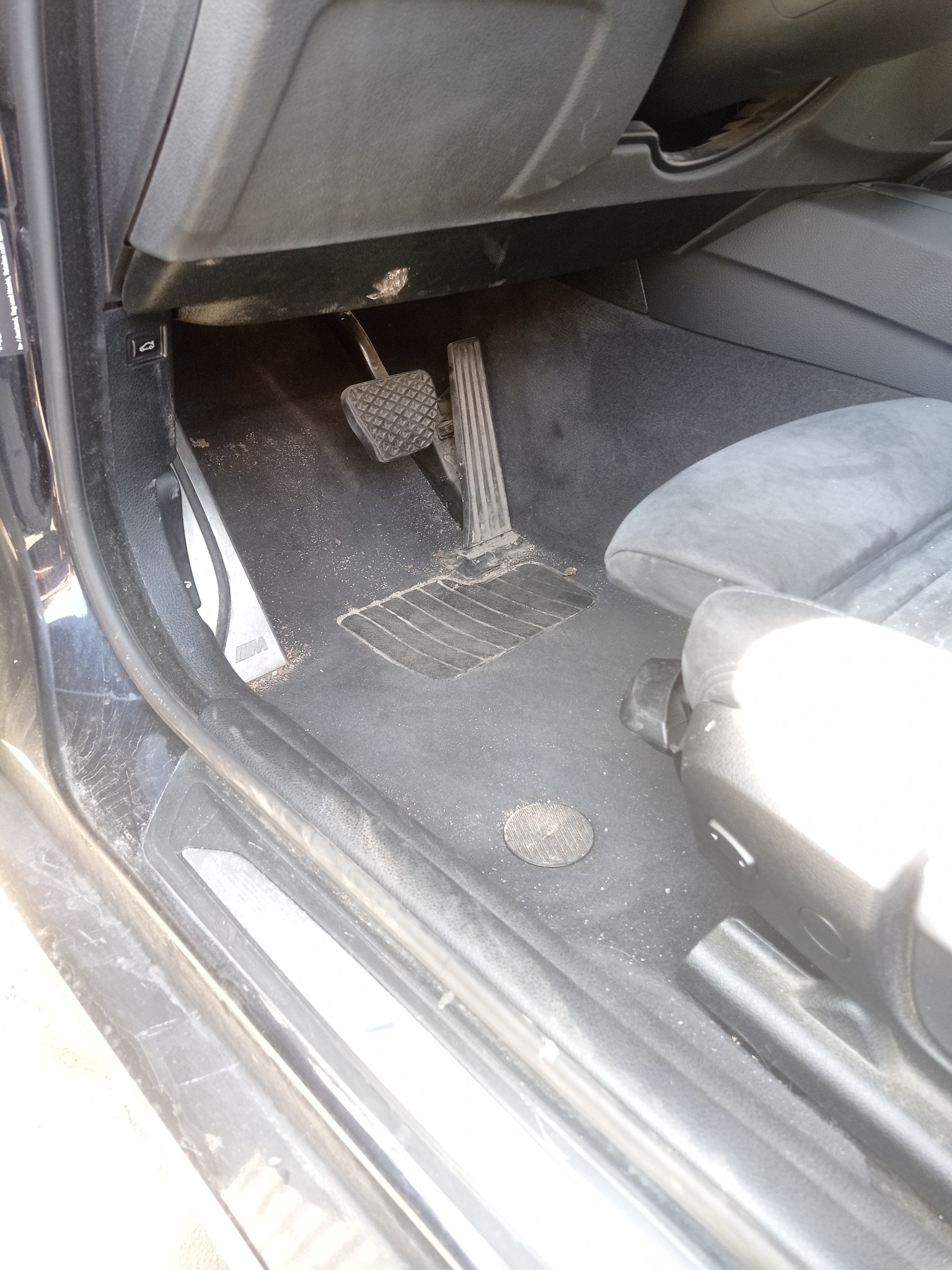 Brudna tapicerka podłogowa i fotel samochodowy w ciemnym wnętrzu pojazdu, widok z dołu na pedały. Wnętrze wymaga odkurzenia i czyszczenia.