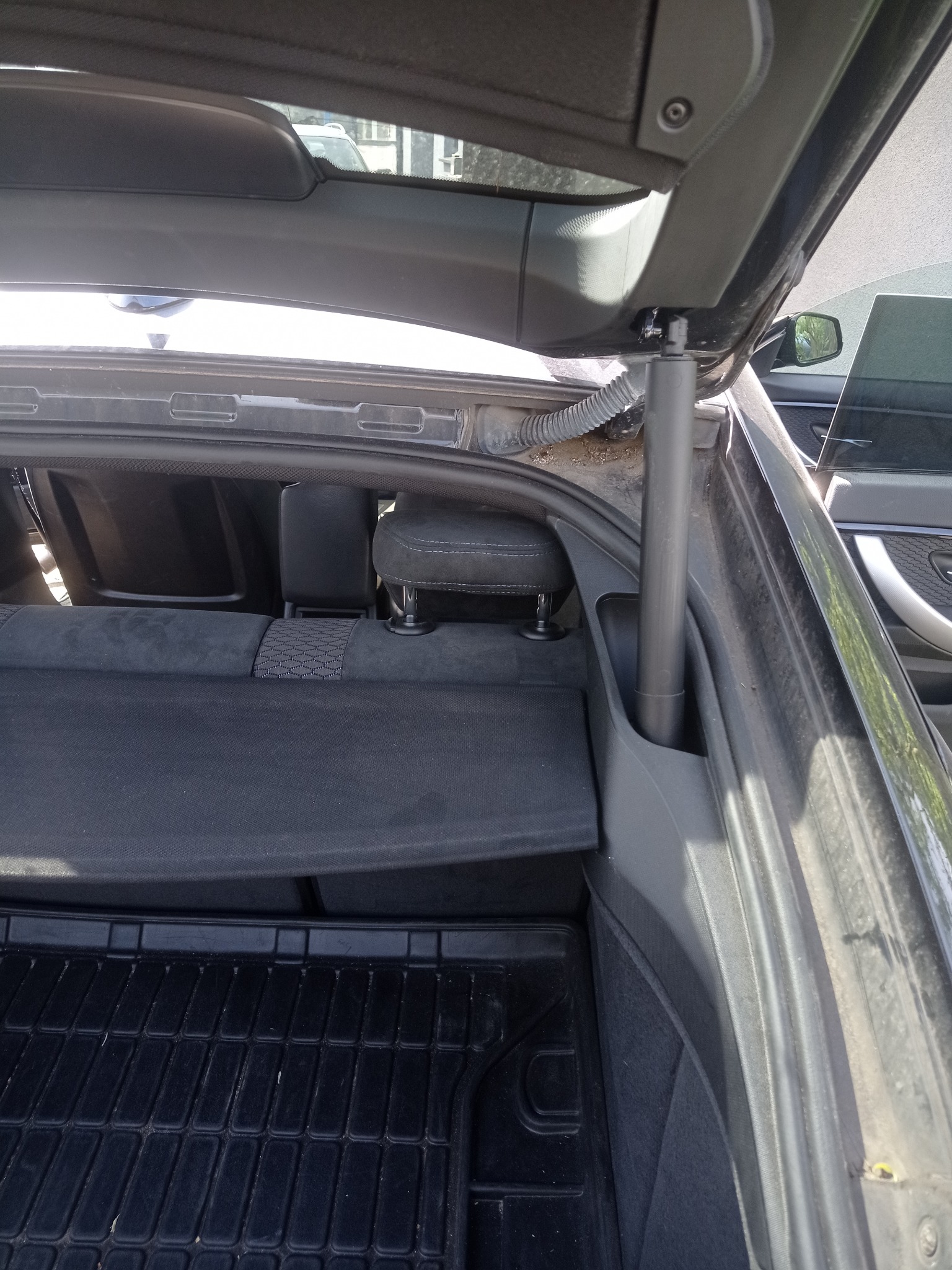 Otwarty bagażnik czarnego samochodu z widoczną tapicerką, gumową wycieraczką i elementami mocowania klapy bagażnika. Widoczne zabrudzenia na elementach tapicerki.