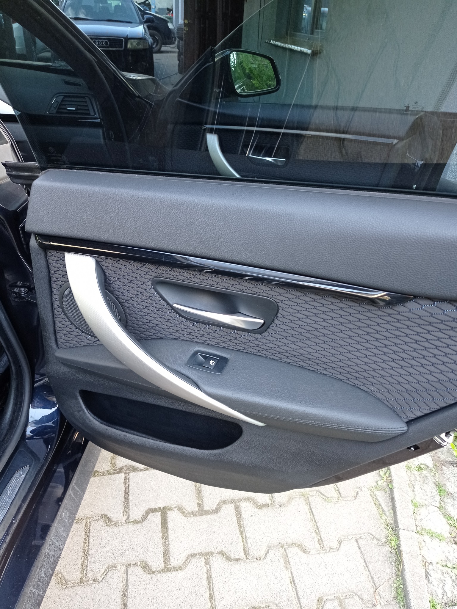 Szczegółowe ujęcie wnętrza samochodu: tapicerka drzwi z srebrną klamką i wzorzystym panelem. Widoczny fragment szyby i karoserii w ciemnym kolorze.