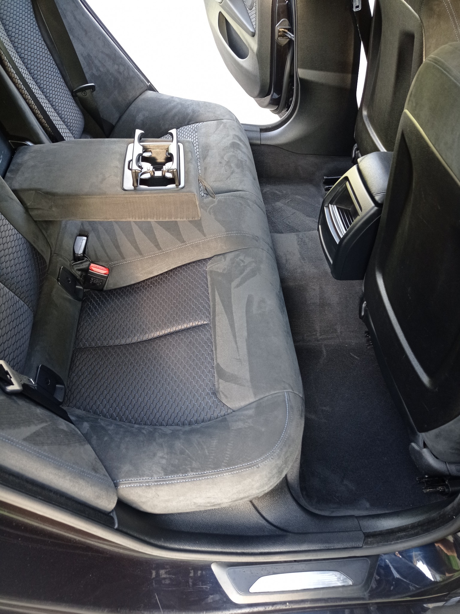 Wnętrze samochodu z szarą tapicerką. Widoczne tylne siedzenia z podłokietnikiem i uchwytami na napoje. Dywaniki wymagają czyszczenia, widoczne zabrudzenia.