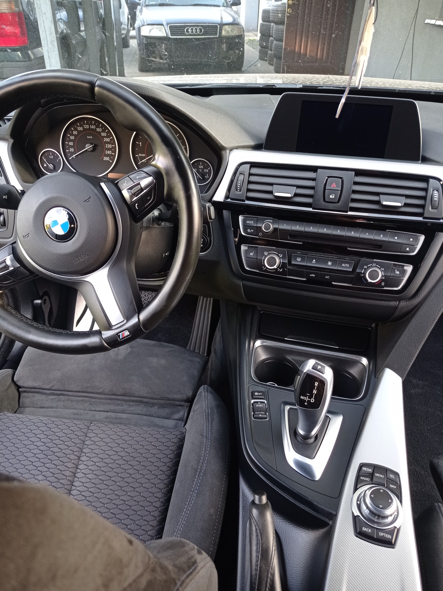 Wnętrze BMW serii 3: kierownica M, czarna tapicerka z wzorem, deska rozdzielcza z ekranem i srebrnymi akcentami, widok na szybę i fragment innego auta.