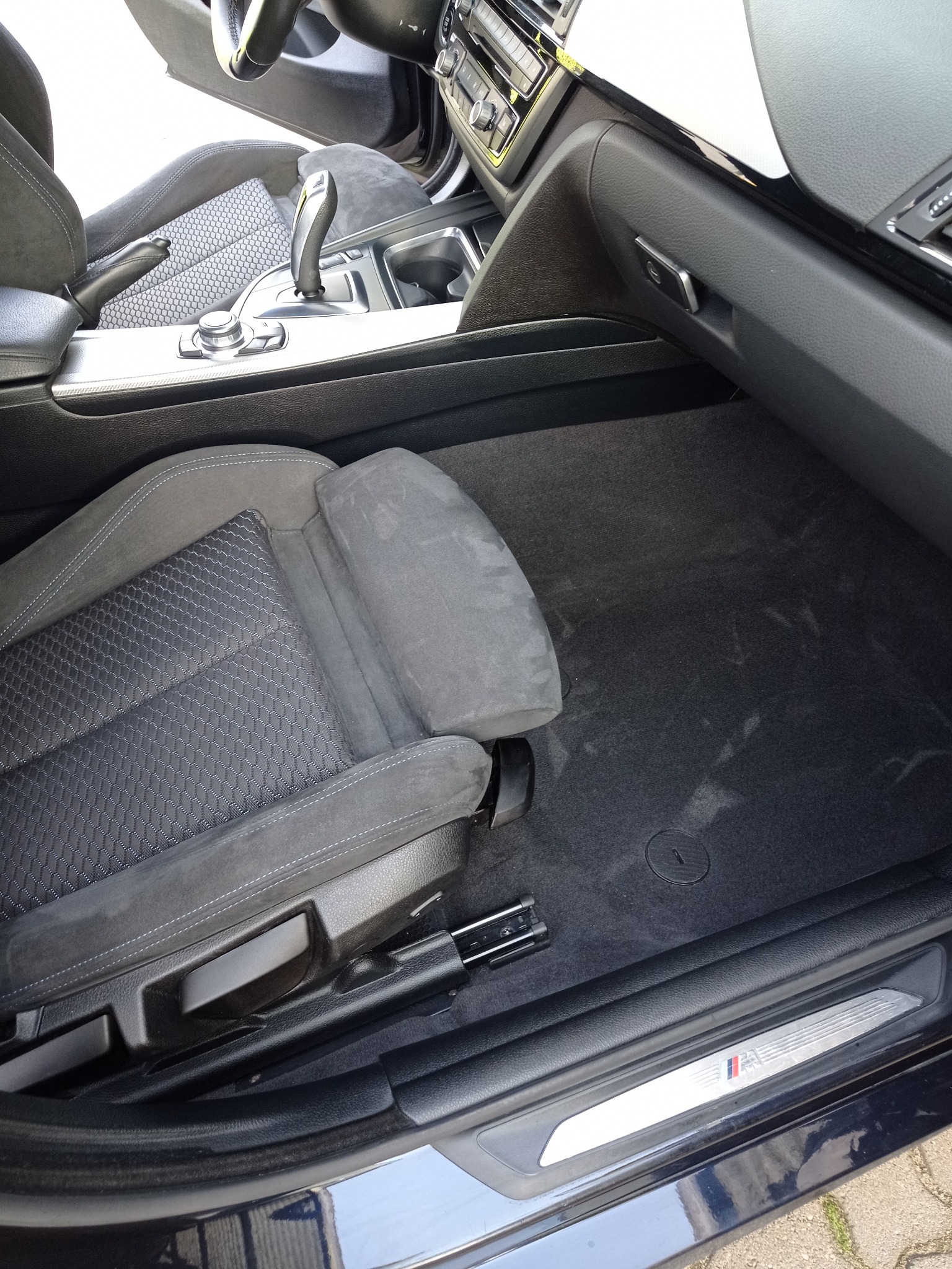 Wnętrze samochodu marki BMW z widocznym fotelem kierowcy, czarną wykładziną podłogową z lekkimi śladami użytkowania i elementami deski rozdzielczej.