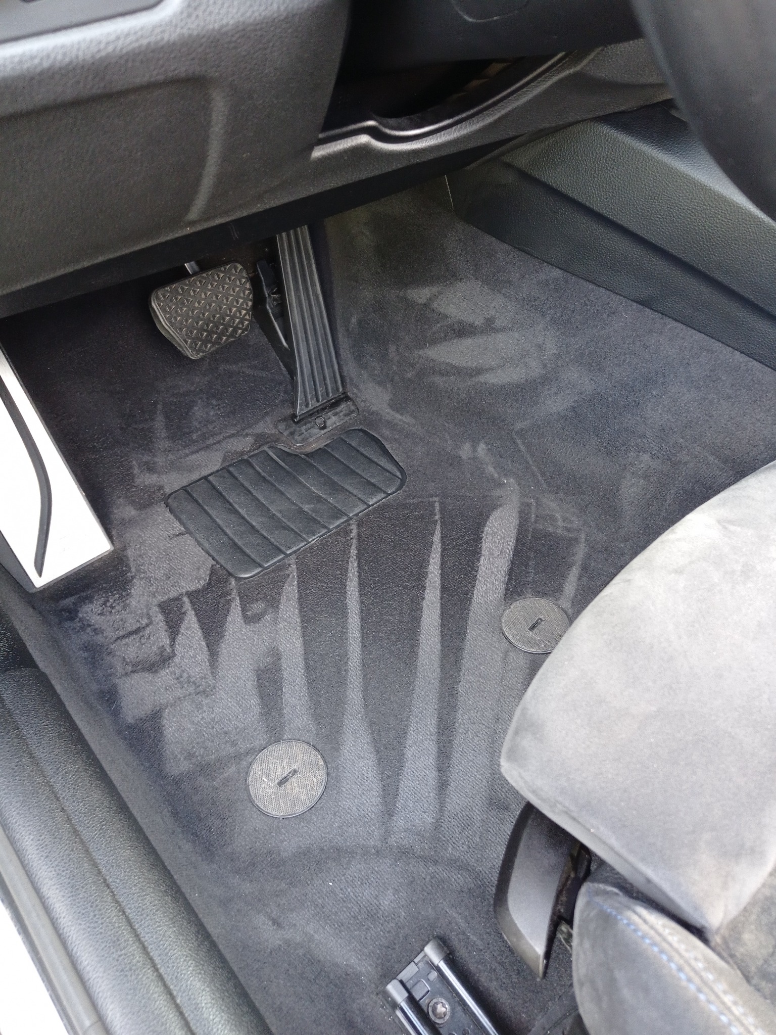 Wnętrze auta po czyszczeniu: świeże ślady po praniu na dywaniku pod pedałami i w okolicy fotela. Ciemna tapicerka, widoczne mocowania dywanika.