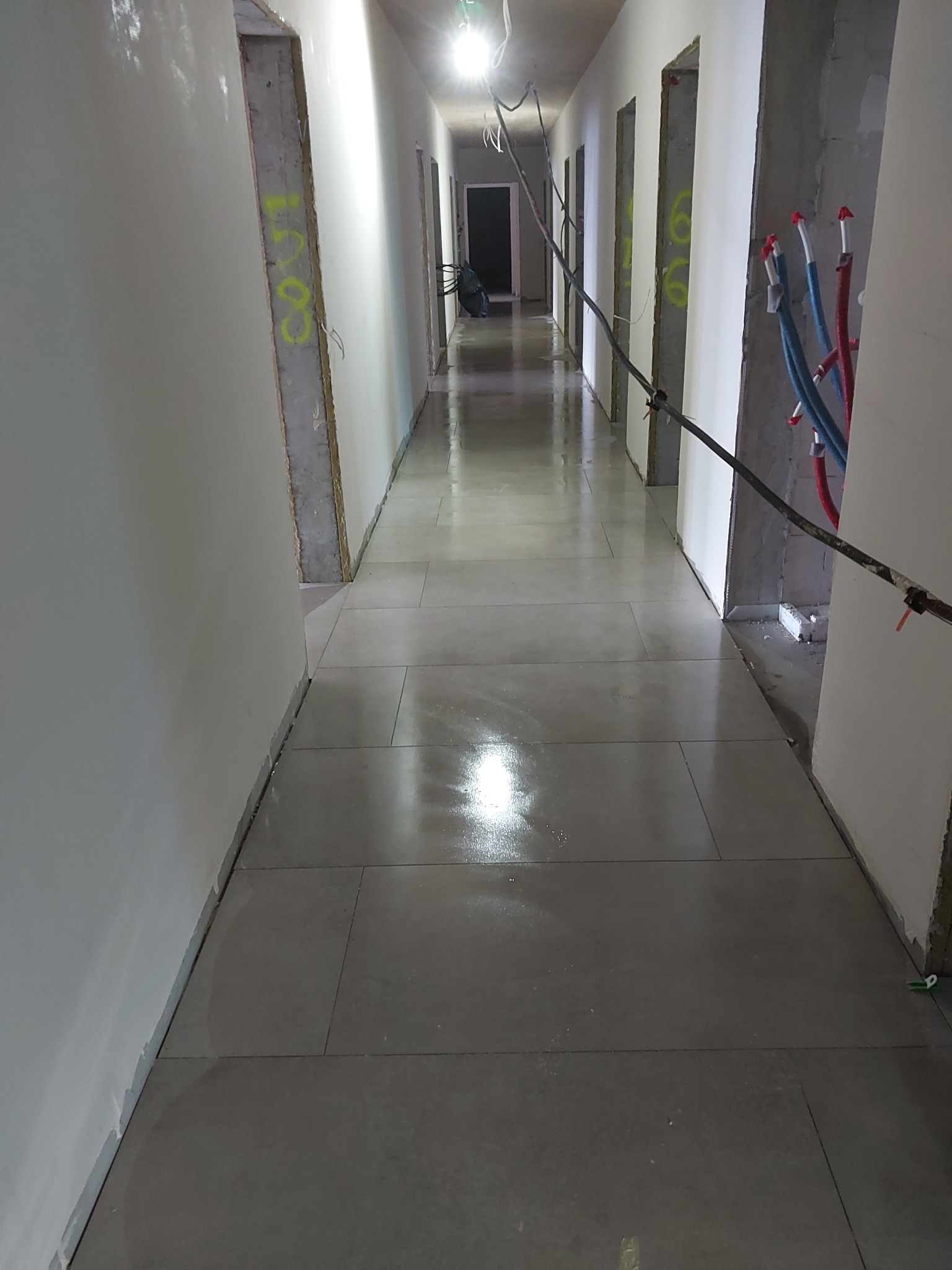 Długi korytarz w trakcie wykańczania: świeżo położone, błyszczące, szare płytki podłogowe, otynkowane ściany z widocznymi instalacjami i surowymi framugami drzwi.