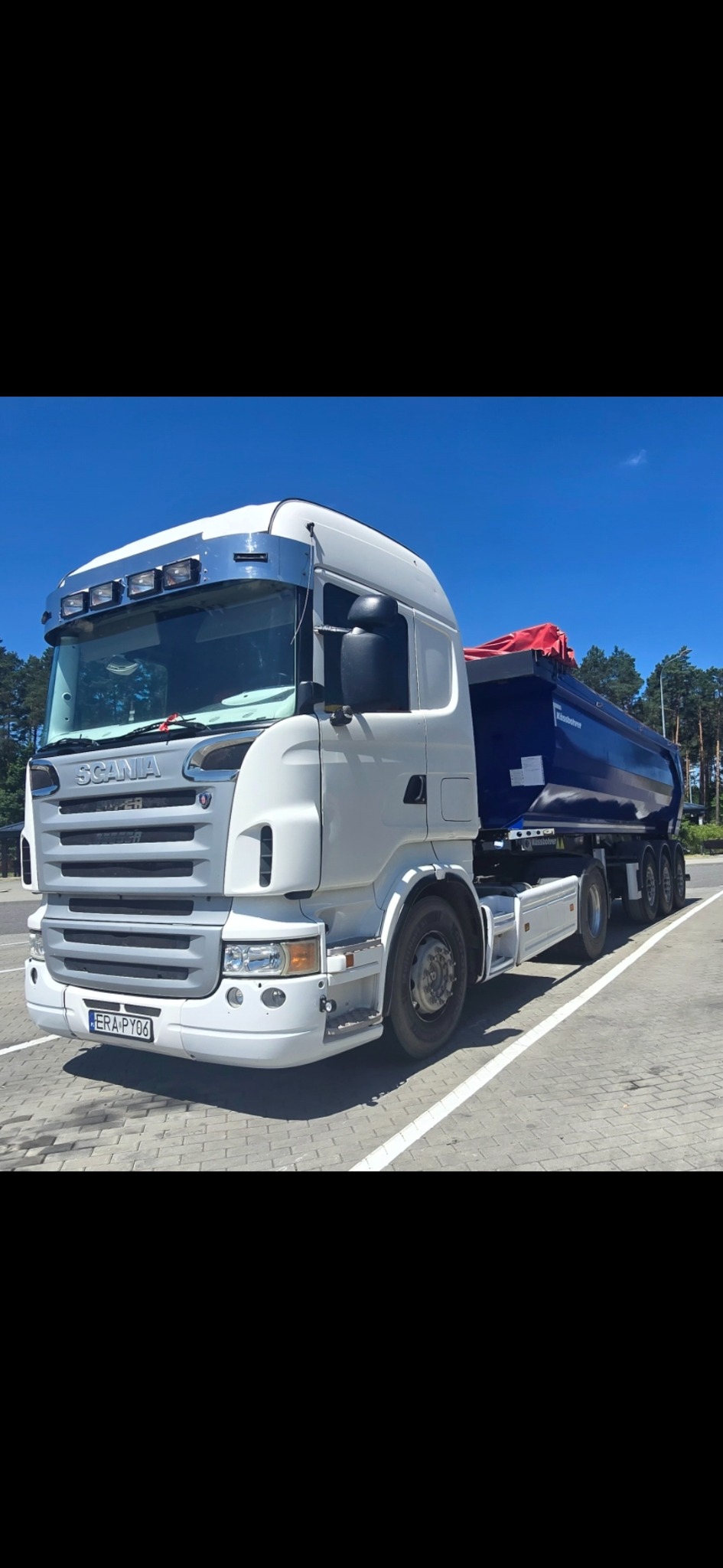 Biała ciężarówka Scania z niebieską naczepą na parkingu, widok z boku, błękitne niebo w tle. Naczepa częściowo zakryta czerwoną plandeką.