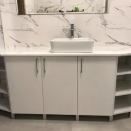 Biała szafka łazienkowa pod umywalkę z blatem imitującym marmur, kwadratowa umywalka nablatowa, srebrna bateria, w tle marmurowe płytki.