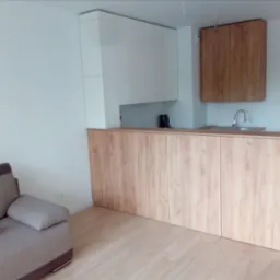 Jasna kuchnia z drewnianym blatem i białymi szafkami, widok z salonu z szarą sofą i kubkiem. Minimalistyczny design w nowoczesnym mieszkaniu.