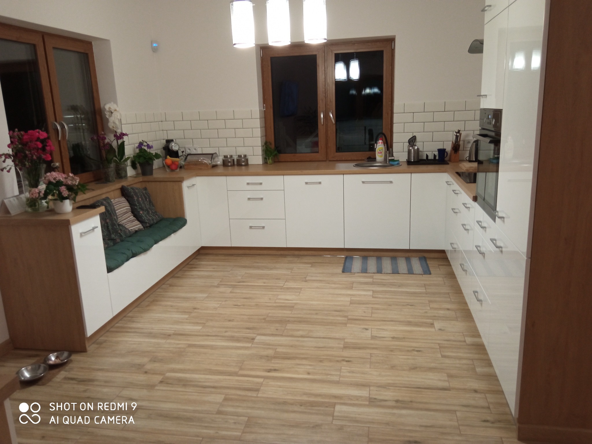 Jasna kuchnia z białymi szafkami, drewnianymi blatami i oknami. W rogu siedzisko z poduszkami. Podłoga imitująca drewno. Nad oknem lampa.