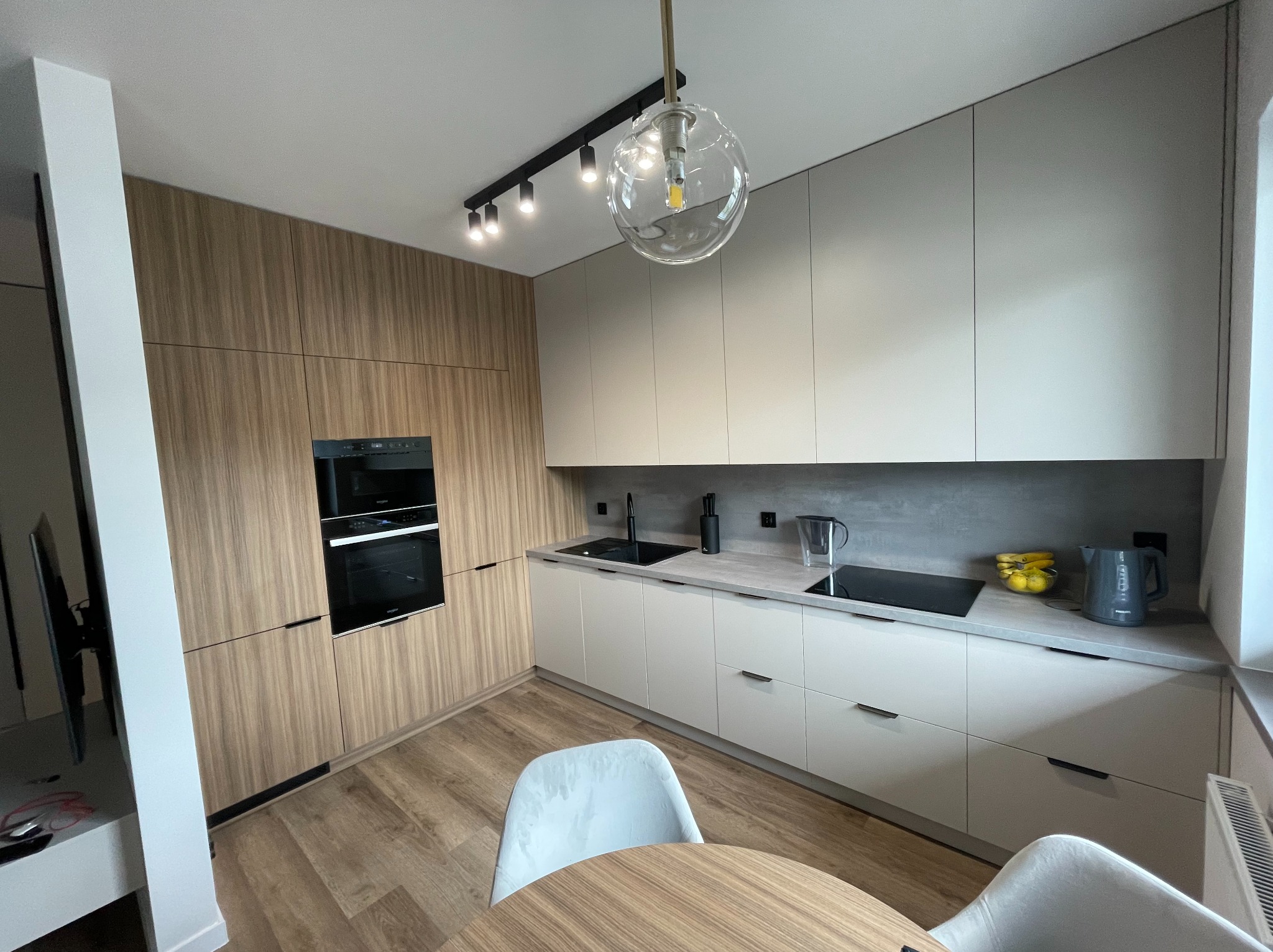 Nowoczesna kuchnia z drewnianymi i szarymi frontami, czarnym zlewem i płytą indukcyjną. Nad blatem lampa wisząca w kształcie kuli. Drewniana podłoga i okrągły stół.