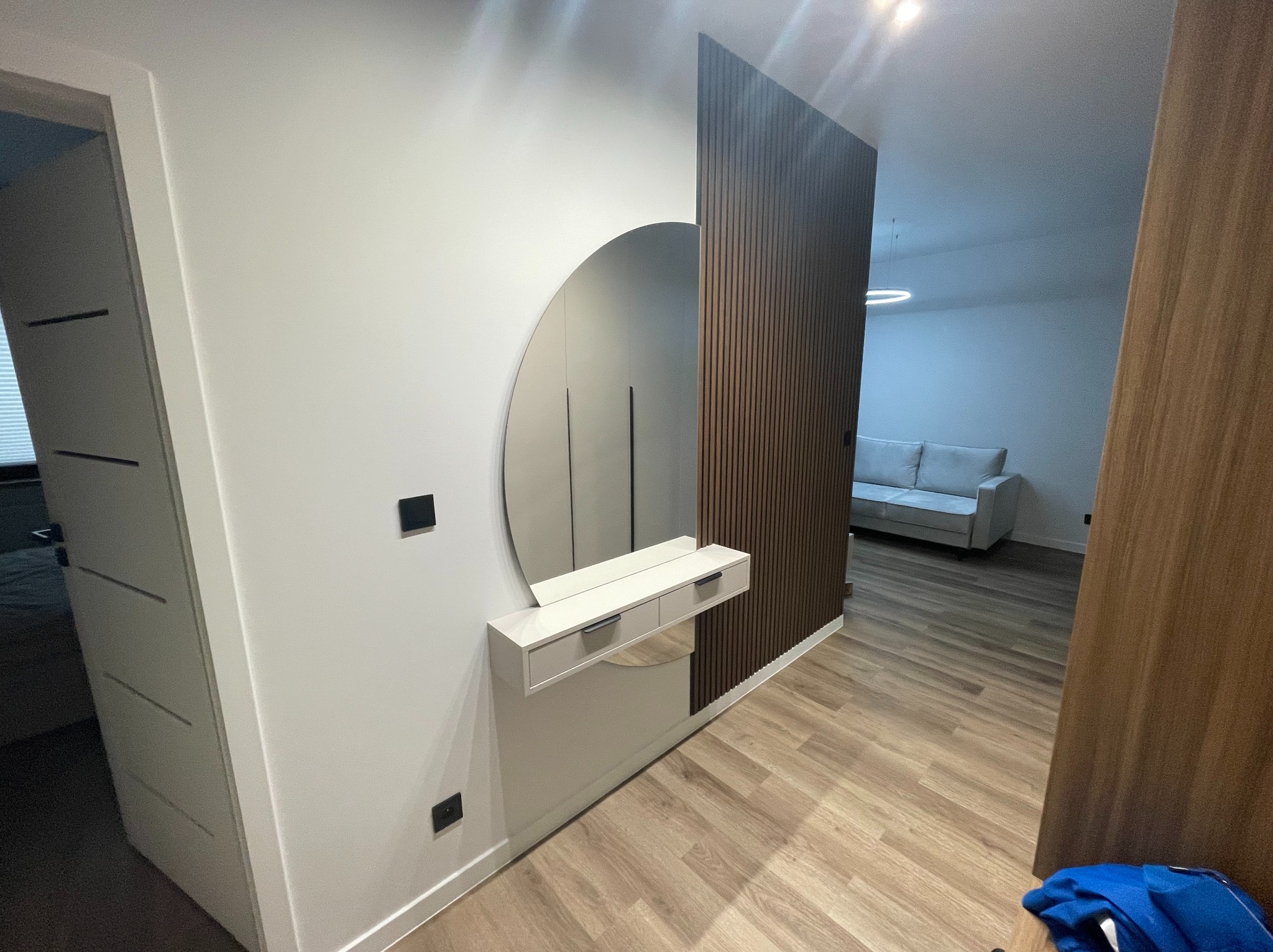 Nowoczesny przedpokój z okrągłym lustrem, konsolą z szufladami i dekoracyjną ścianką z drewnianych listewek. Widok na salon z sofą. Jasne drewno na podłodze.